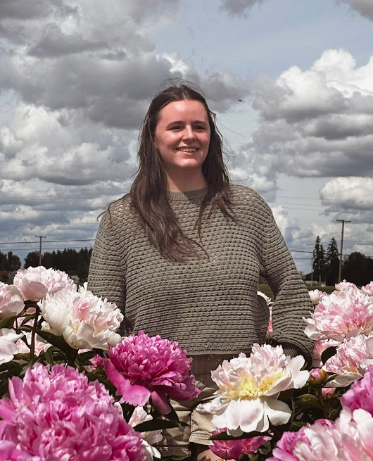 A young woman smiling outdoors among blooming peonies under a cloudy sky.