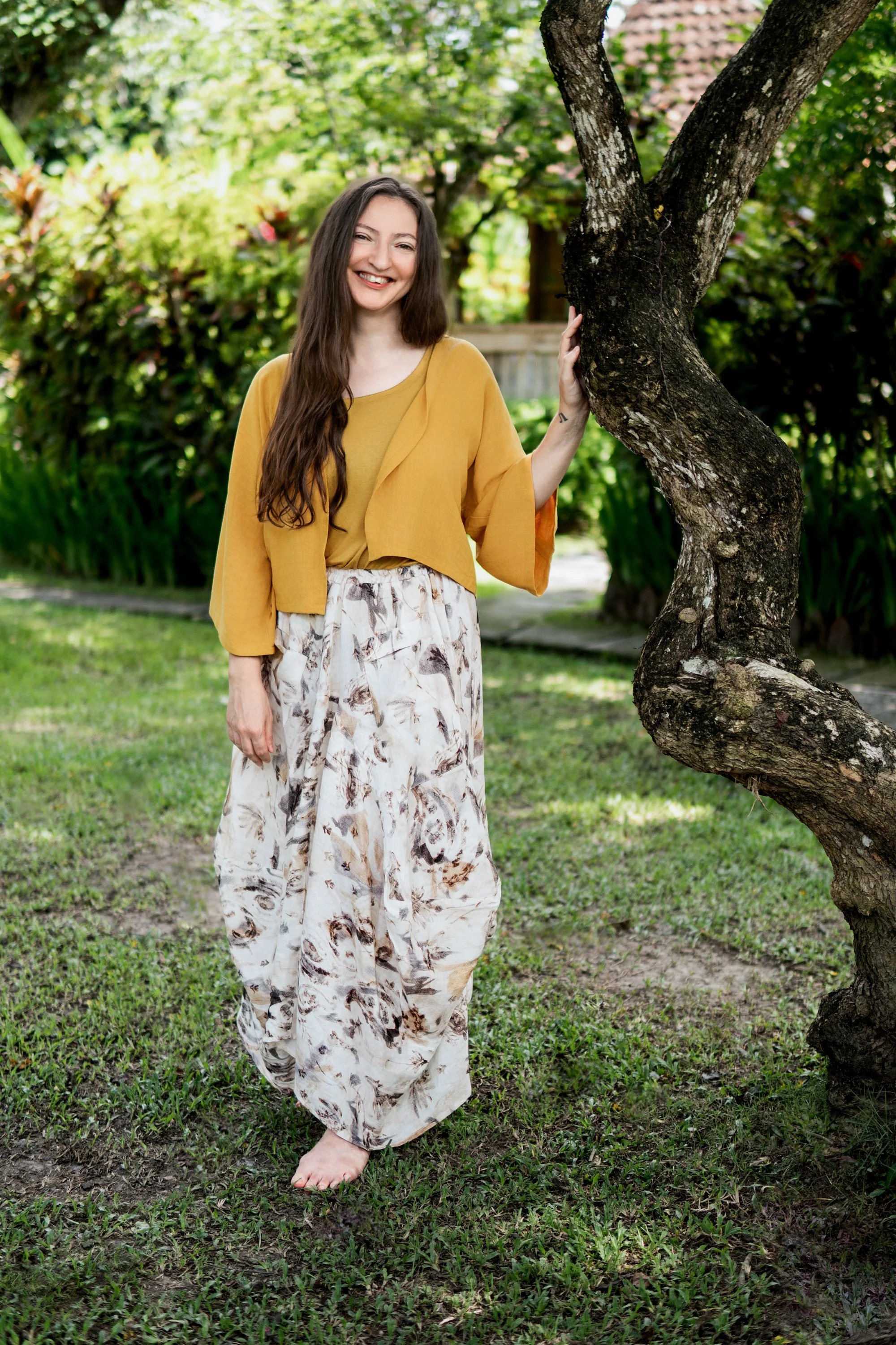 A young woman with long brown hair standing outdoors near a tree, smiling and wearing a yellow top and wide patterned pants, with one hand touching the tree.