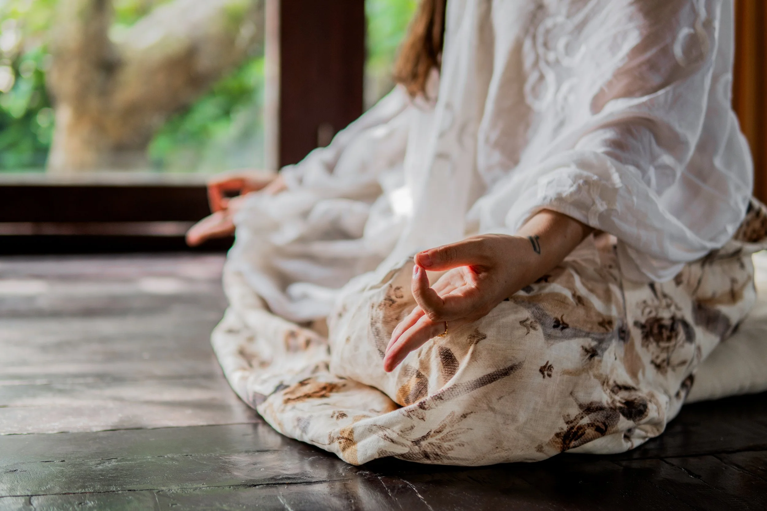 A person sitting cross-legged on what appears to be a wooden floor, practicing meditation or yoga indoors, with a view of greenery outside through a window.