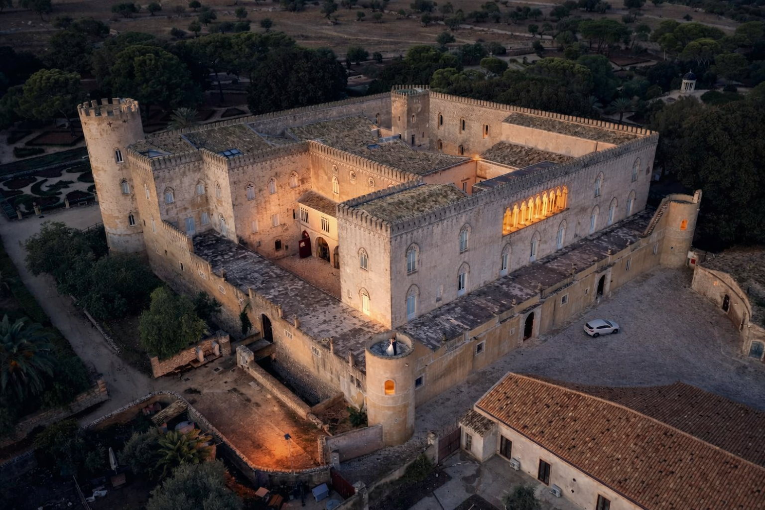 Aerial view of a large historic castle with stone walls and towers, lit up at dusk, surrounded by greenery and some buildings.