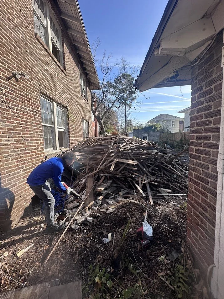 A person wearing a blue jacket and gray pants appears to be working with tools near a large pile of broken wooden debris between two brick buildings on a clear, sunny day.