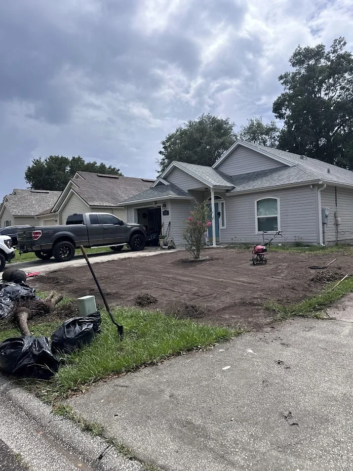 Front yard of a house with the grass and plants removed, indicating yard renovation or landscaping work, with a small pink toy motorcycle on the dirt area, and a tree with pink flowers near the house, under an overcast sky.