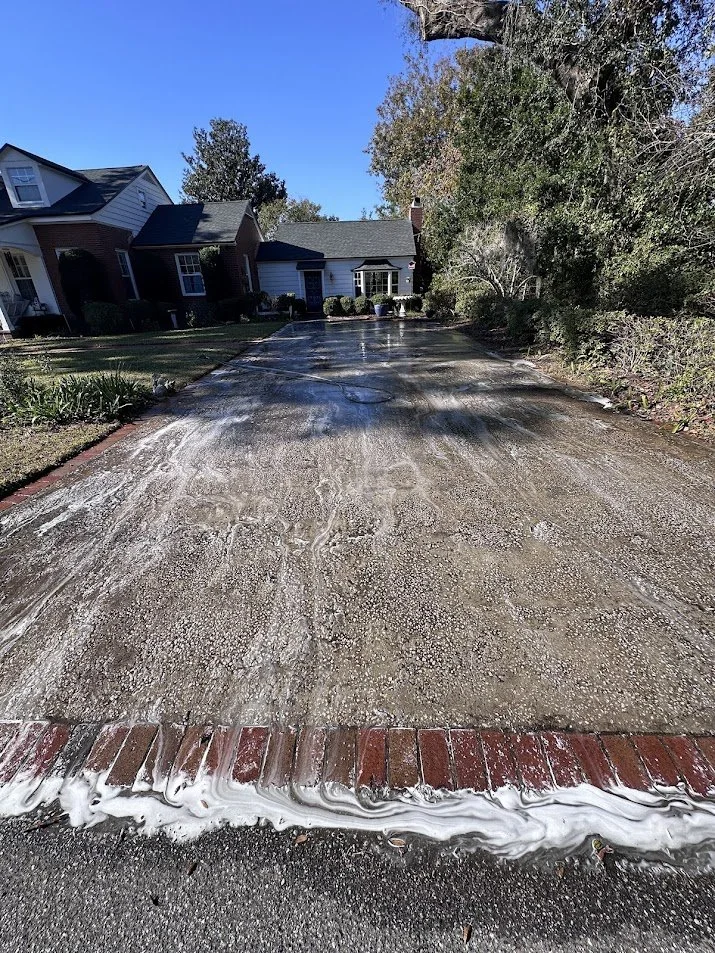 A residential driveway being washed with water, partially covered by a red brick border, with houses and trees in the background under a clear blue sky.