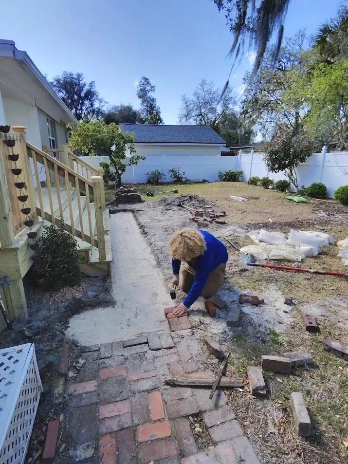 A person is laying bricks to build a new walkway in their backyard, with a partially finished brick path and tools nearby.