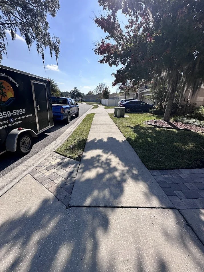 Sidewalk on a residential street with a shadow of a tree, parked cars, a landscaping trailer, and front yards with grass and trees.