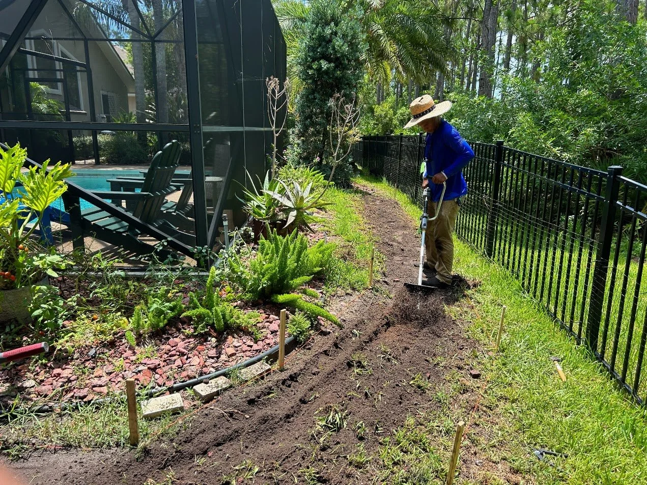 Person wearing a wide-brimmed hat and blue shirt using a power garden tool to till or dig soil along a fence line next to a landscaped garden with various plants, near a screened porch or pool area in a backyard.