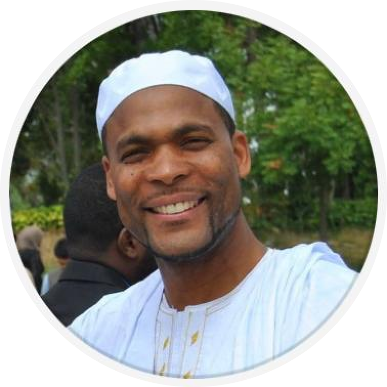 A smiling man wearing a white traditional outfit and cap, outdoors with greenery in the background.
