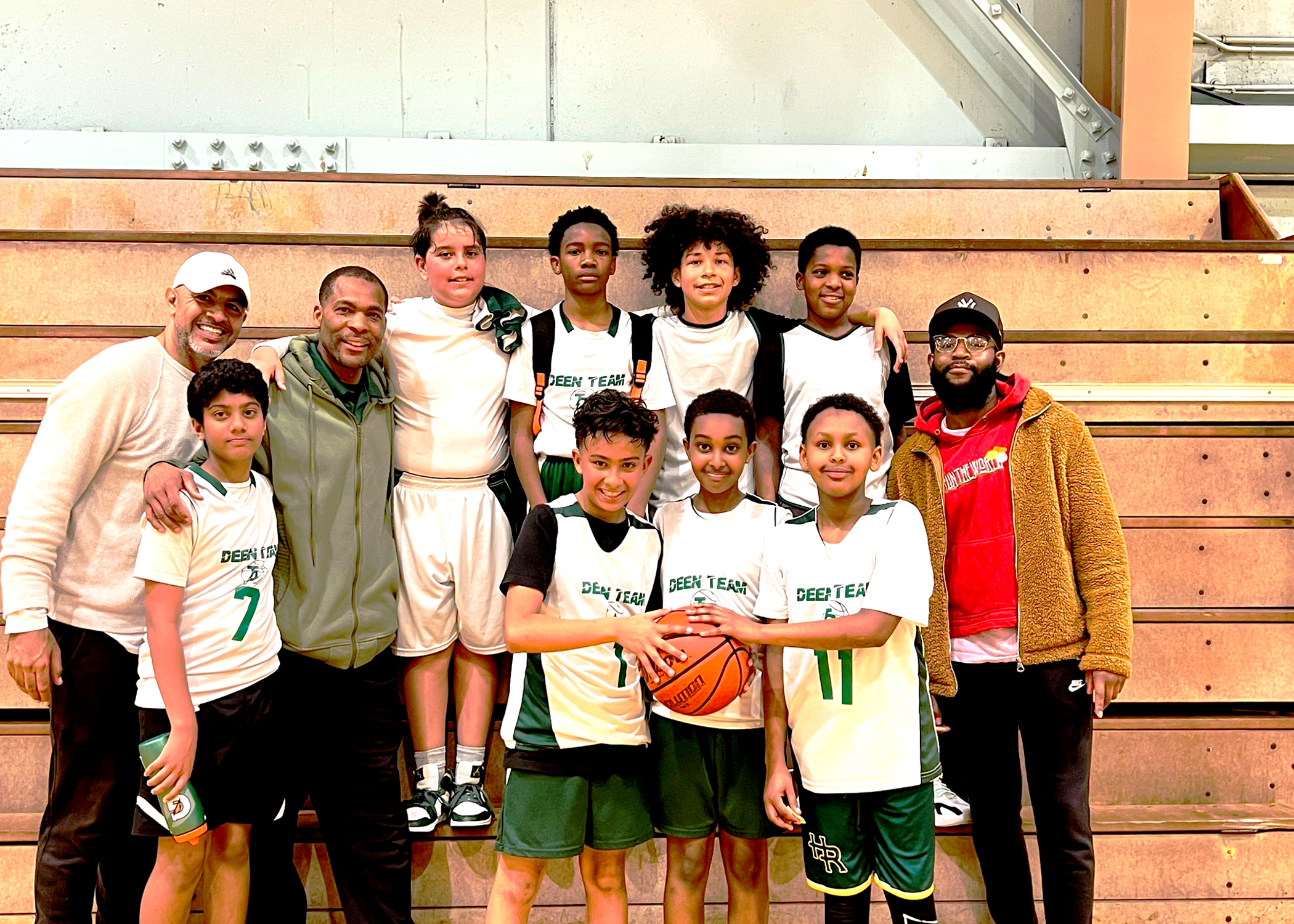 Group of Deen and Basketball young basketball players and coaches posing for a team photo in front of gym bleachers, with some players holding a basketball.
