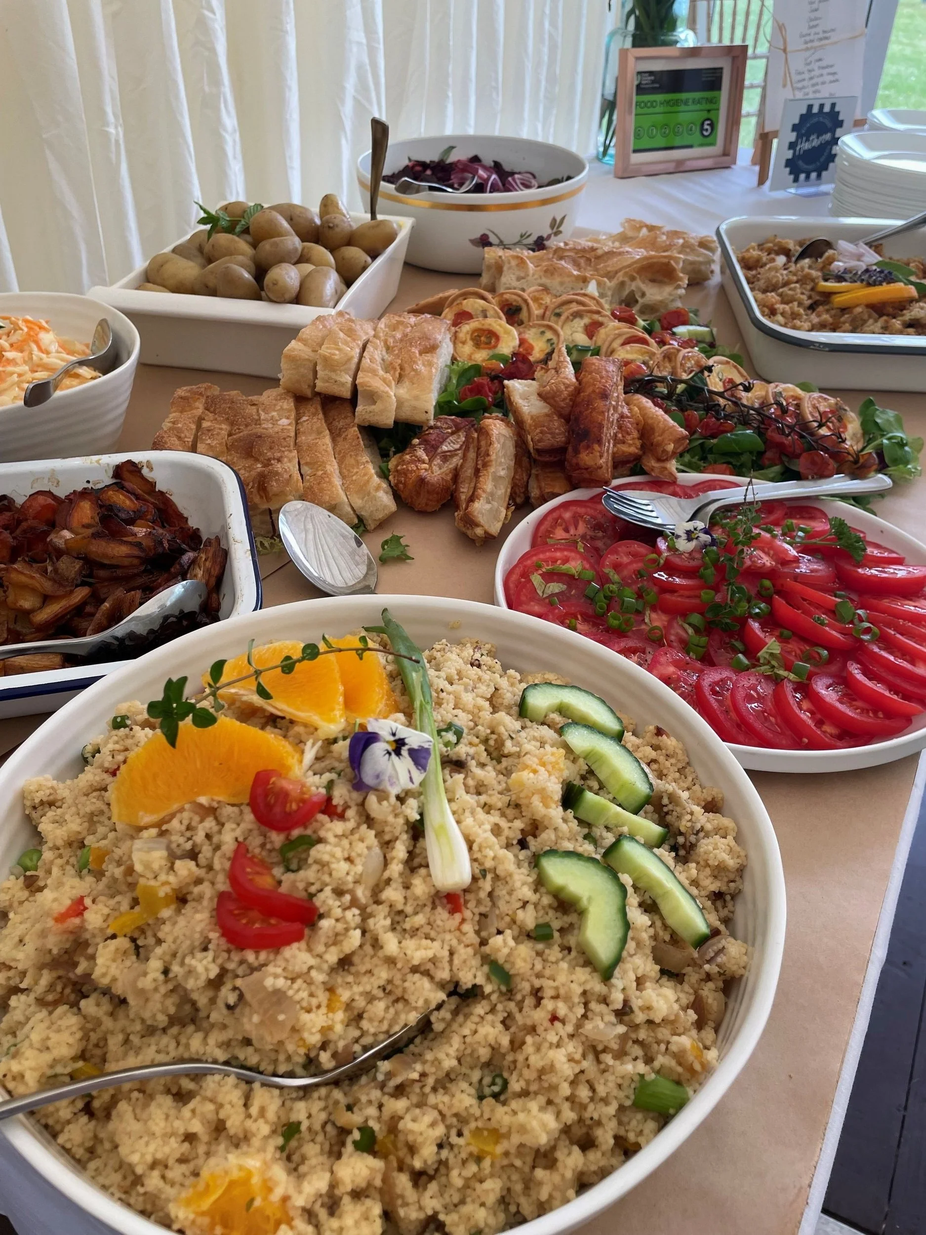 Buffet table with various dishes including couscous salad with cucumbers and tomatoes, red cabbage salad, boiled potatoes, fresh tomato slices, vegetable pastries, and bread slices.