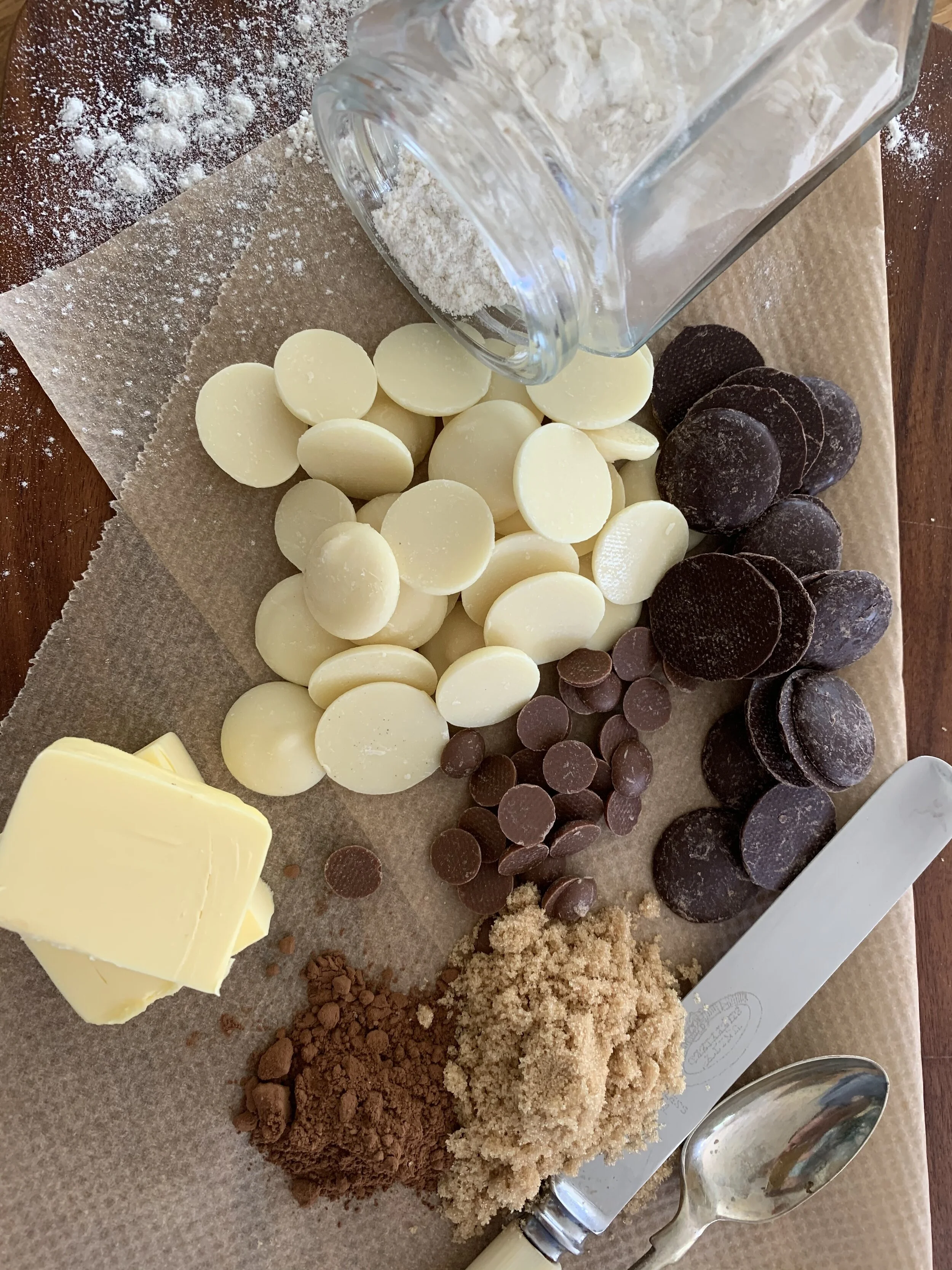 Assorted baking ingredients on parchment, including white and dark chocolate discs, brown sugar, cocoa powder, butter chunks, and a jar of flour. A spoon and knife are nearby.