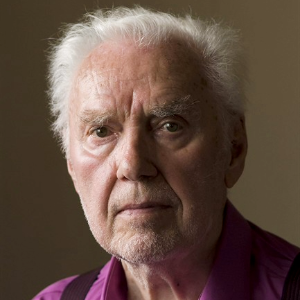 Close-up portrait of an elderly man with white hair and beard wearing a purple shirt against a dark background.