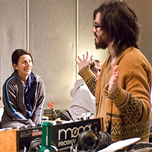 A man with long hair and glasses wearing a brown sweater is speaking with two women at a music event or signing. One woman has dark hair and is wearing a dark jacket, and the other has light hair and glasses. There are musical equipment and items on the table.