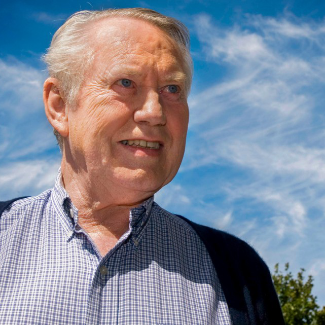 Close-up of an elderly man with white hair wearing a checkered shirt and a dark jacket, smiling outdoors against a blue sky with some clouds.