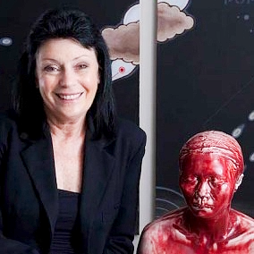 A woman with black hair smiling sitting next to a red sculpture of a person's head.