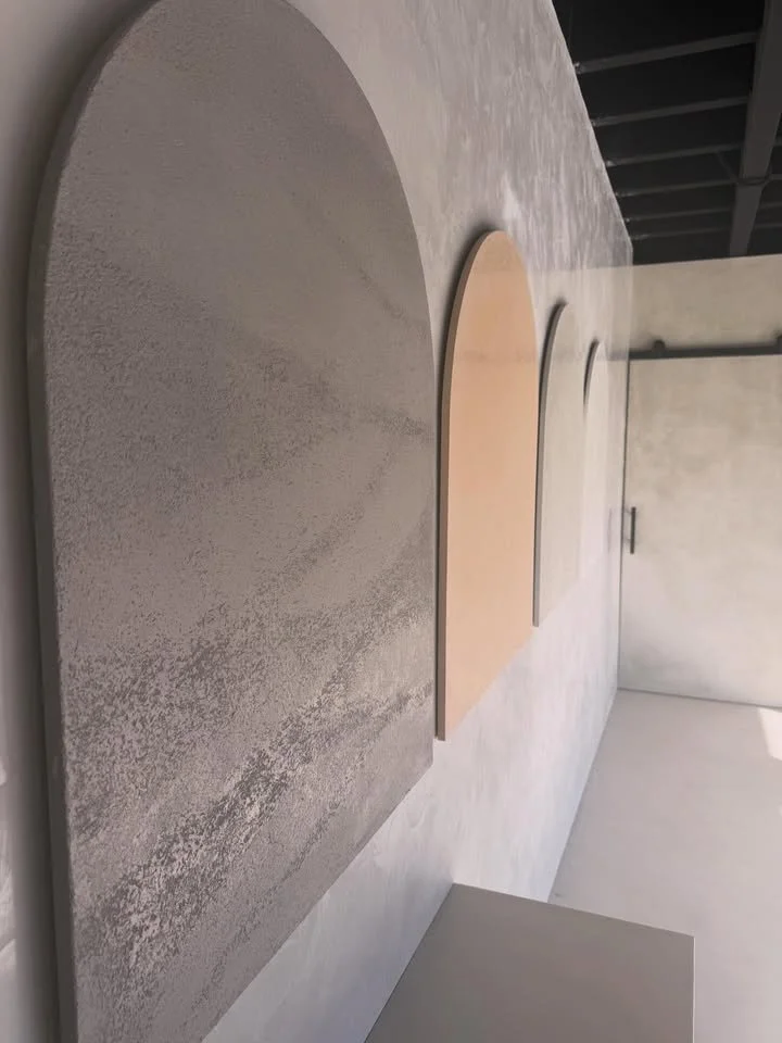 Three semi-circular wall hangings with textured surfaces in gray, beige, and black, mounted on a white wall in a modern interior space. Mornington Peninsula Venetian Plaster showroom