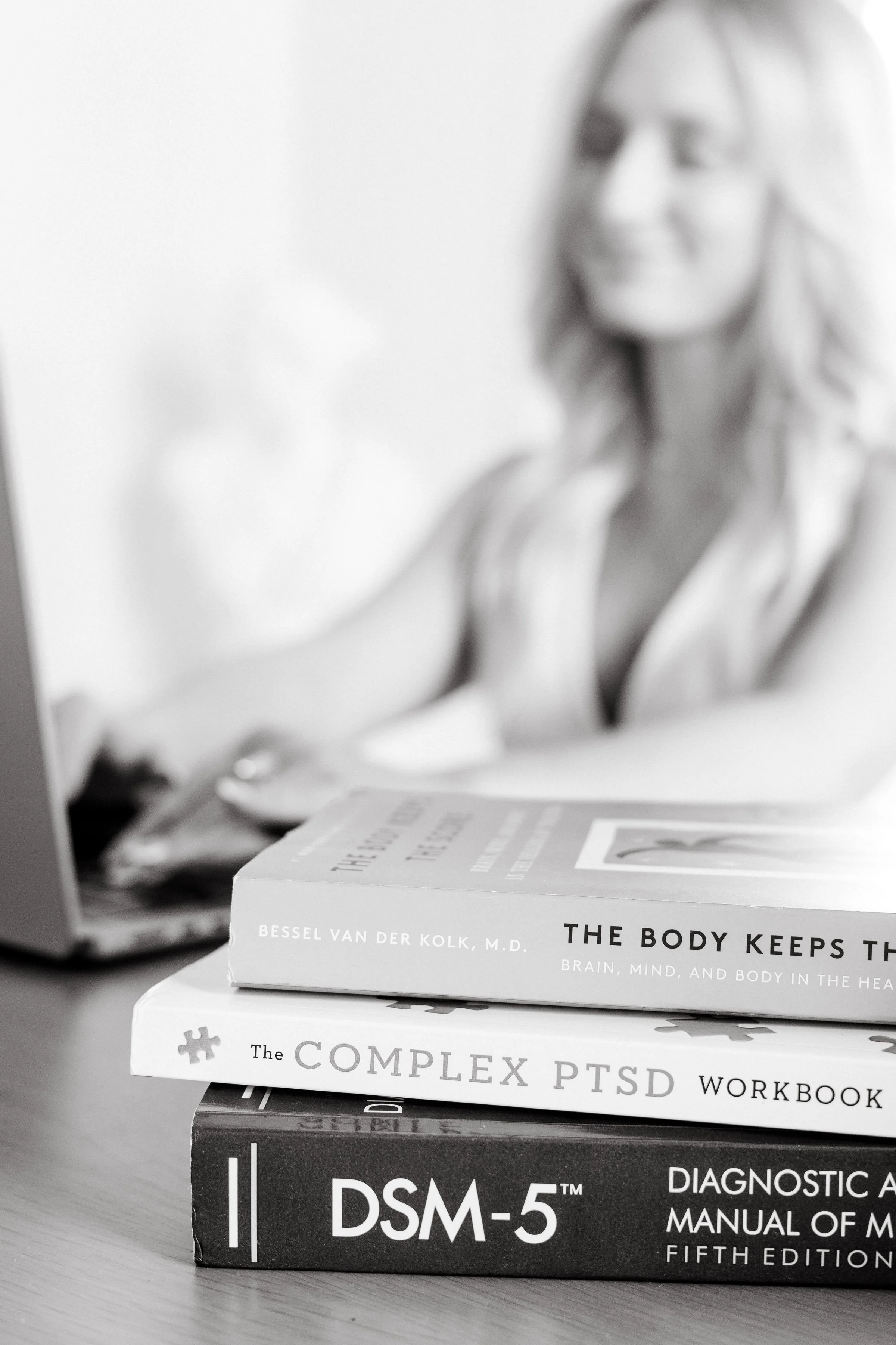 Stack of three books on a desk with a blurred woman in the background on a computer screen.