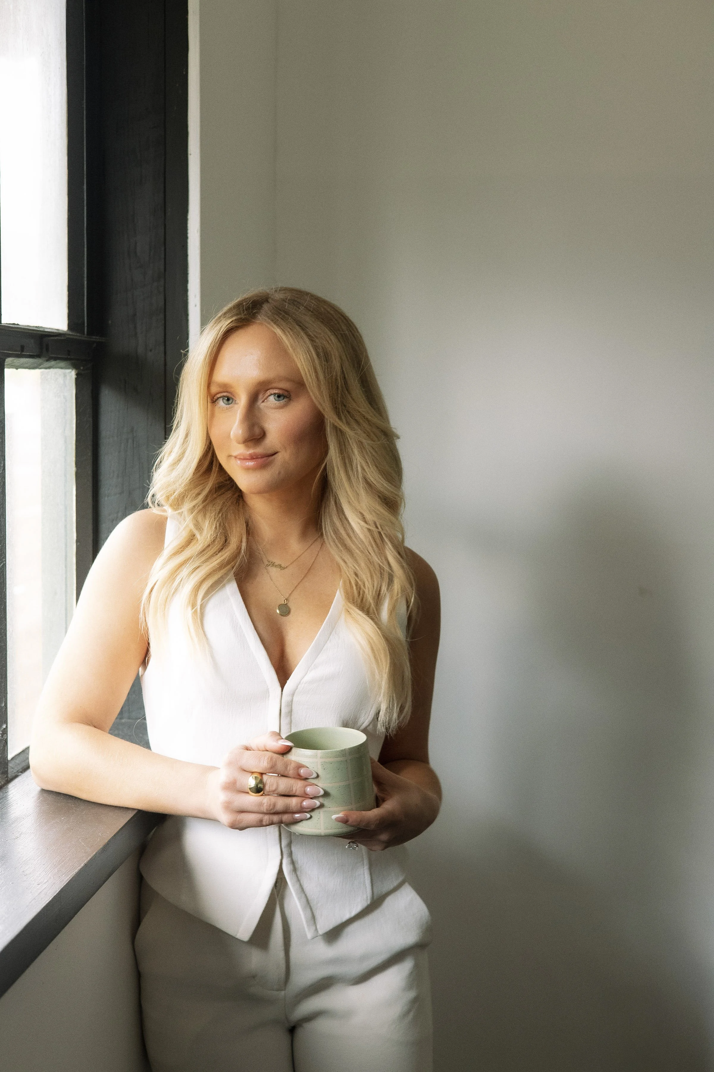 A woman with long blonde hair holding a mug, standing near a window, wearing a white sleeveless top, jewelry, and light-colored pants.