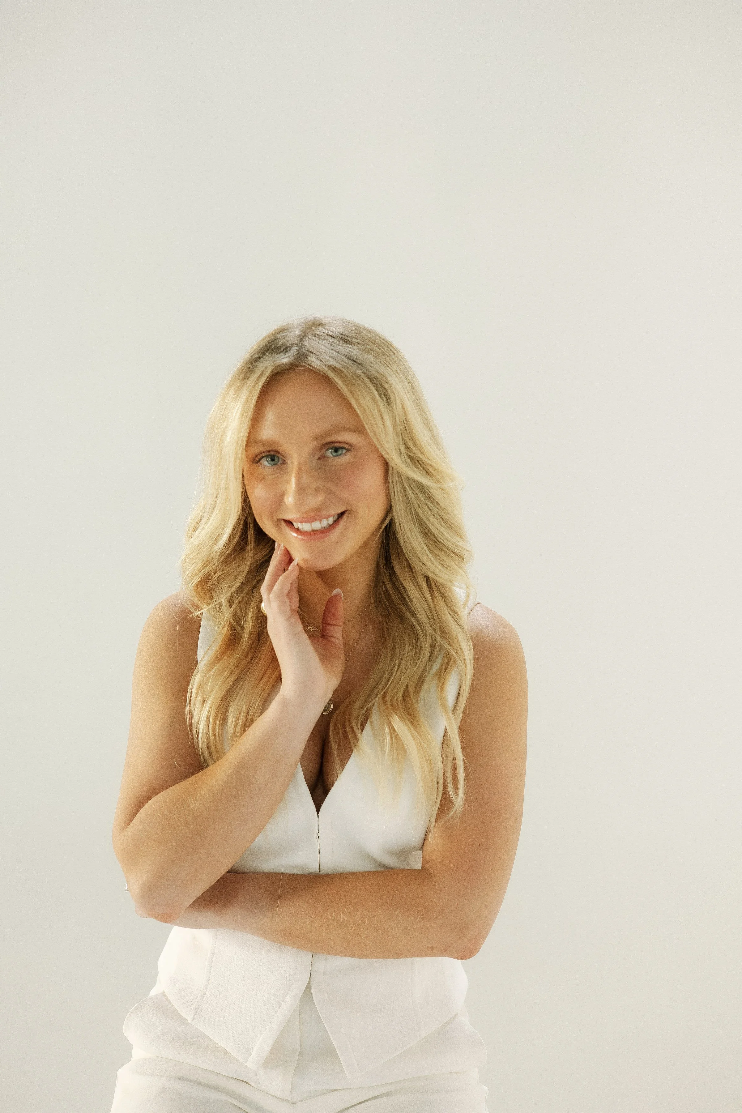 A woman with long blonde hair smiling, wearing a white sleeveless top, and touching her chin with her hand, posing against a plain, light-colored background.