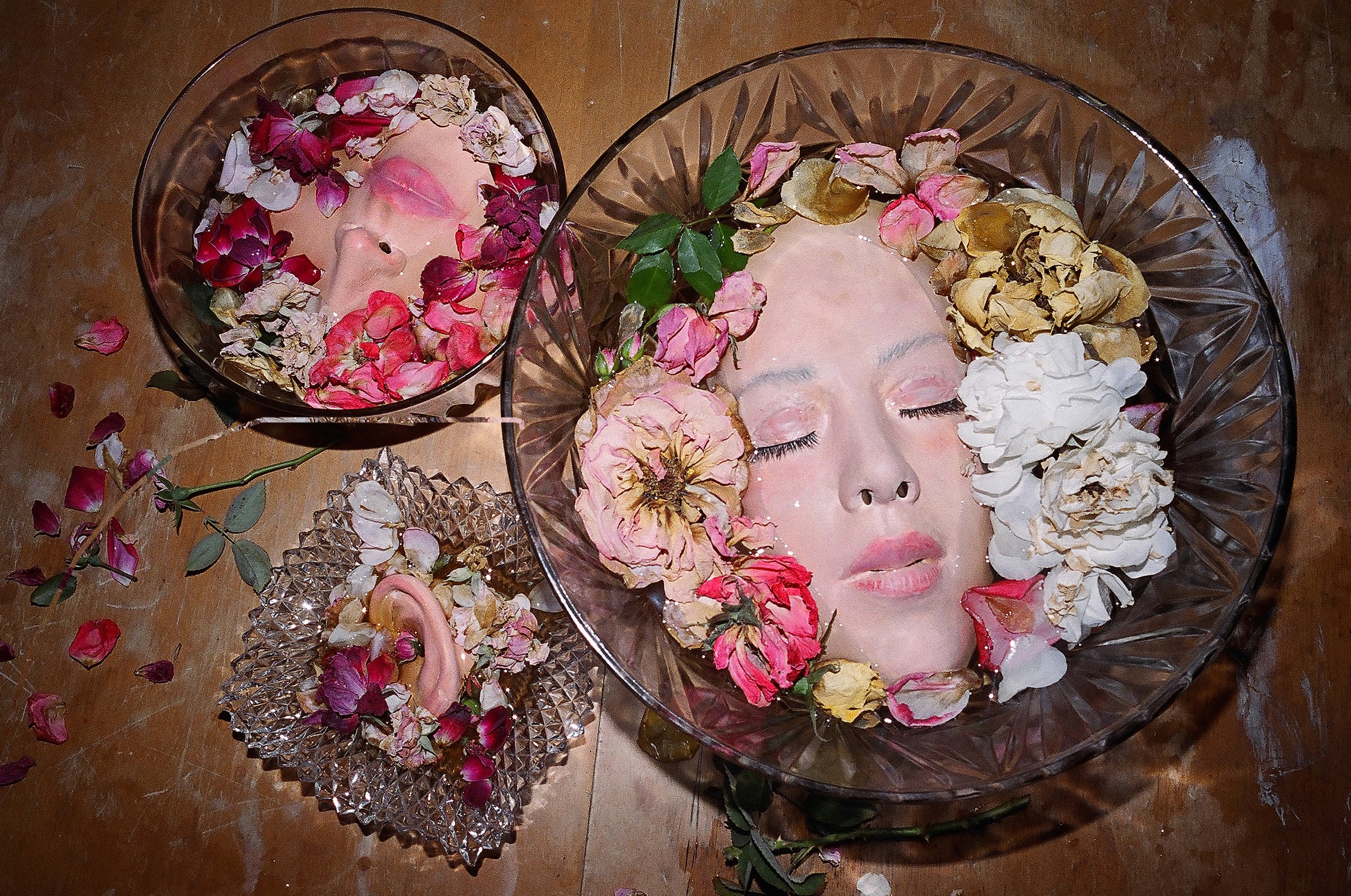 Faces of women submerged in water, surrounded by flower petals in glass bowls.