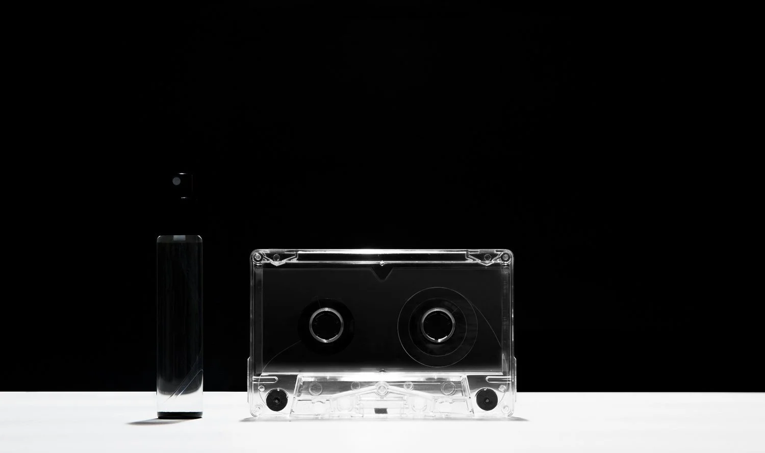 A black and white photo of a small perfume bottle and a transparent cassette tape on a white surface against a black background.