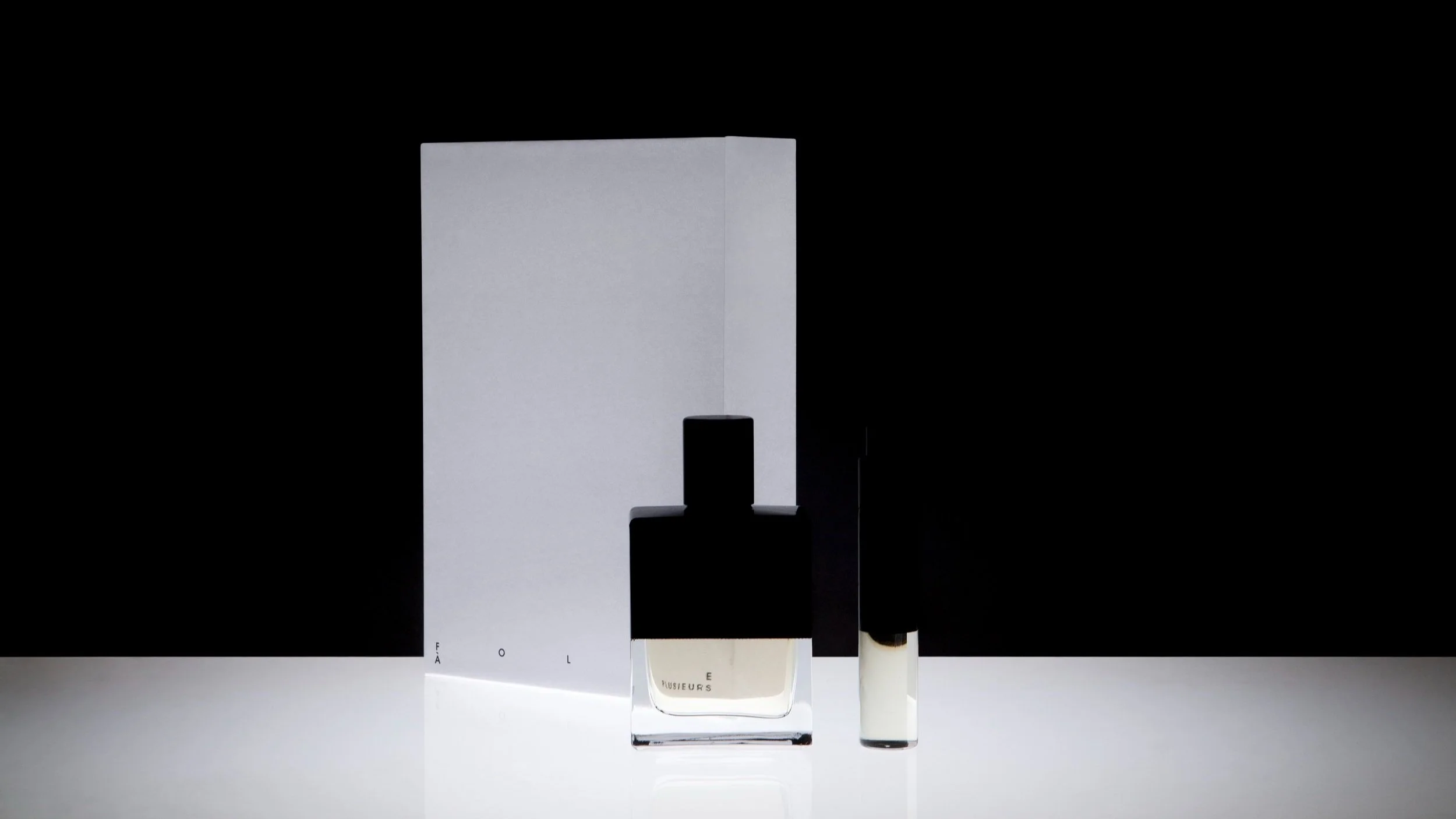 A minimalist perfume bottle with a black cap, a white box in the background, and a small cylindrical container, all set against a black and white background.