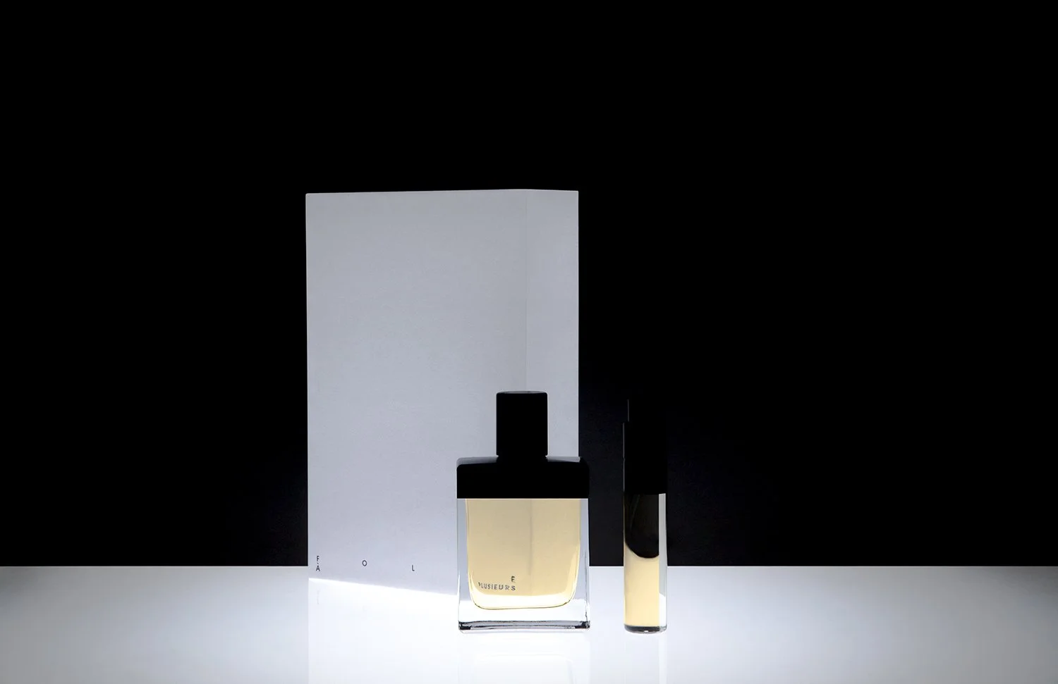 A bottle of perfume and a smaller container on a reflective surface against a black background, with a large light gray book or panel behind them.