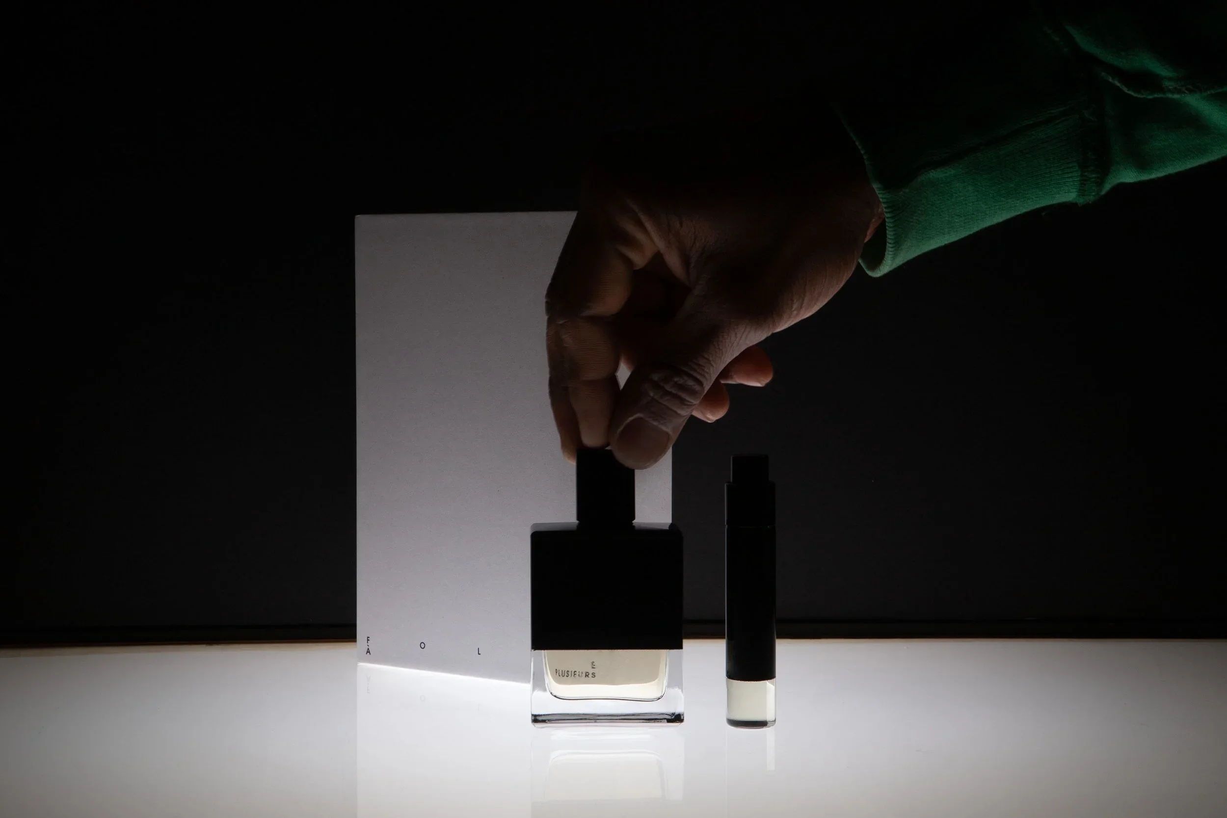 A hand holding a black and transparent bottle of nail polish in front of a dark background and white surface.