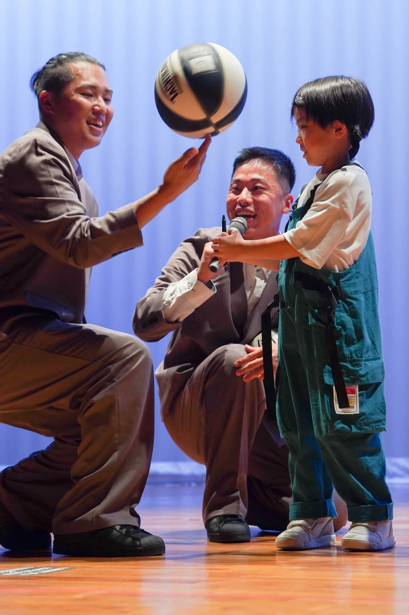 Two adults and a child playing with a volleyball on a stage with a blue background.