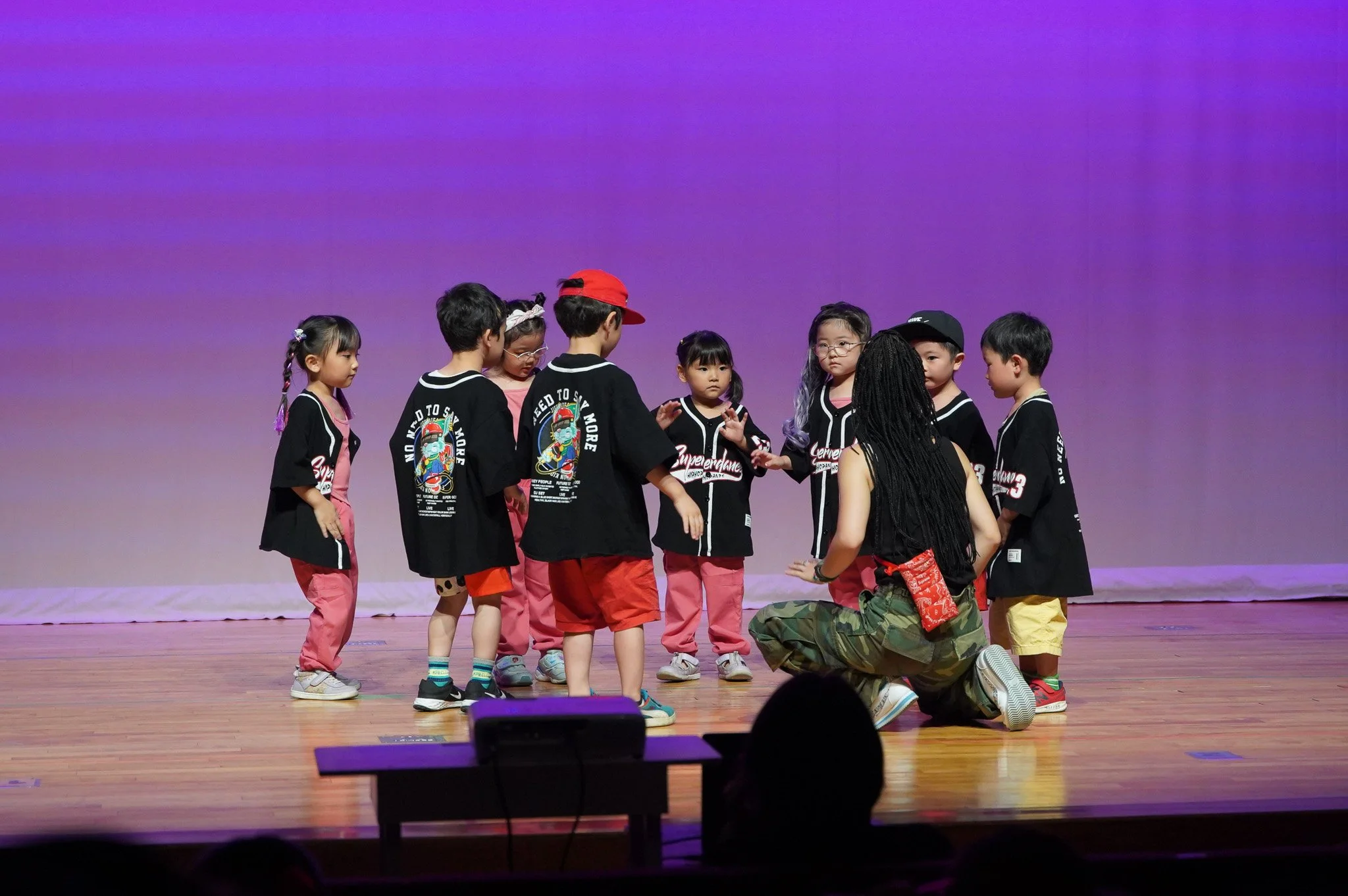 Children on stage participating in a performance or activity, some standing and some kneeling, with a plain purple background.