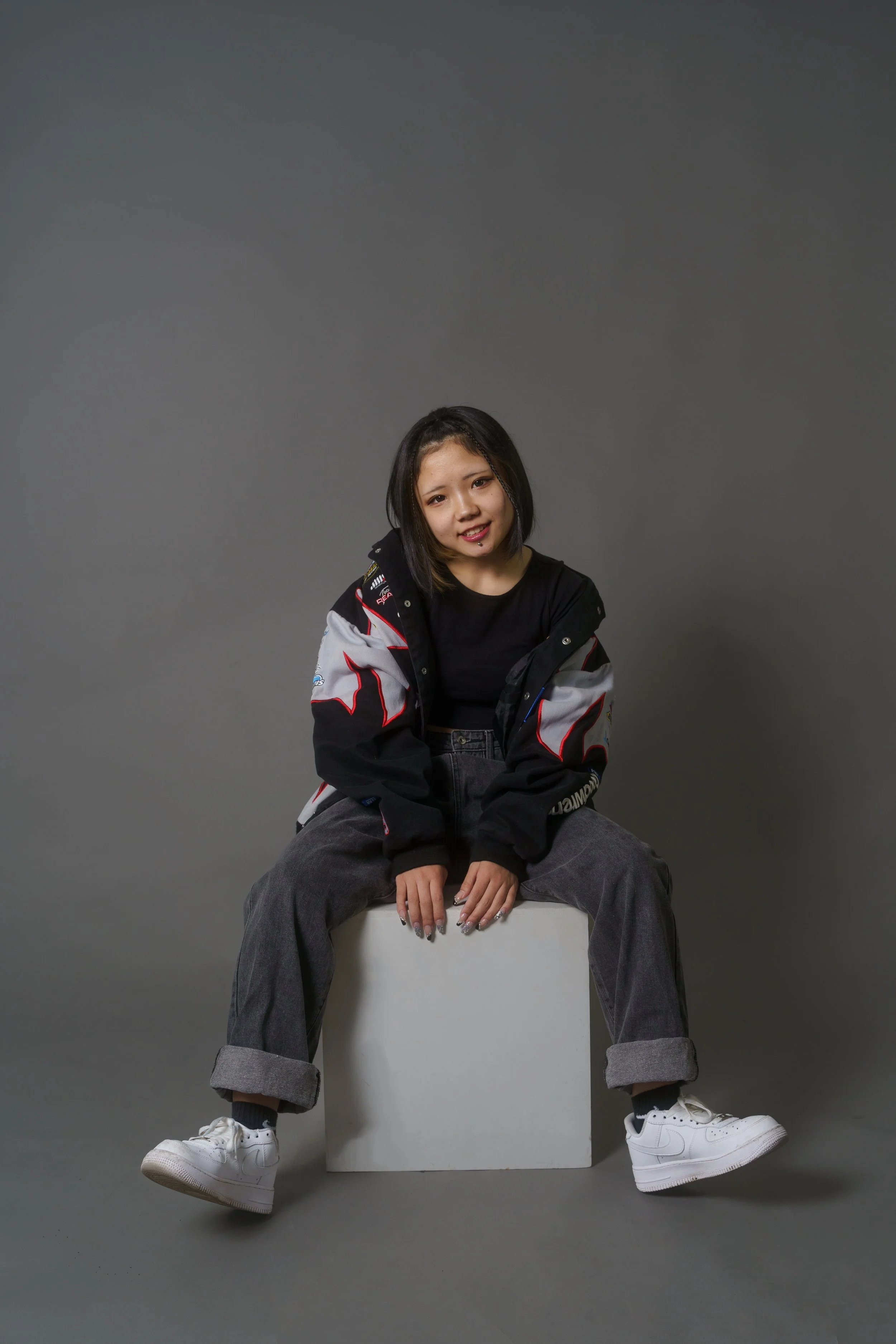 A young woman with black hair sitting on a white cube against a plain gray background, wearing a black T-shirt, gray jeans, a black and gray jacket, white sneakers, and posing with a relaxed expression.