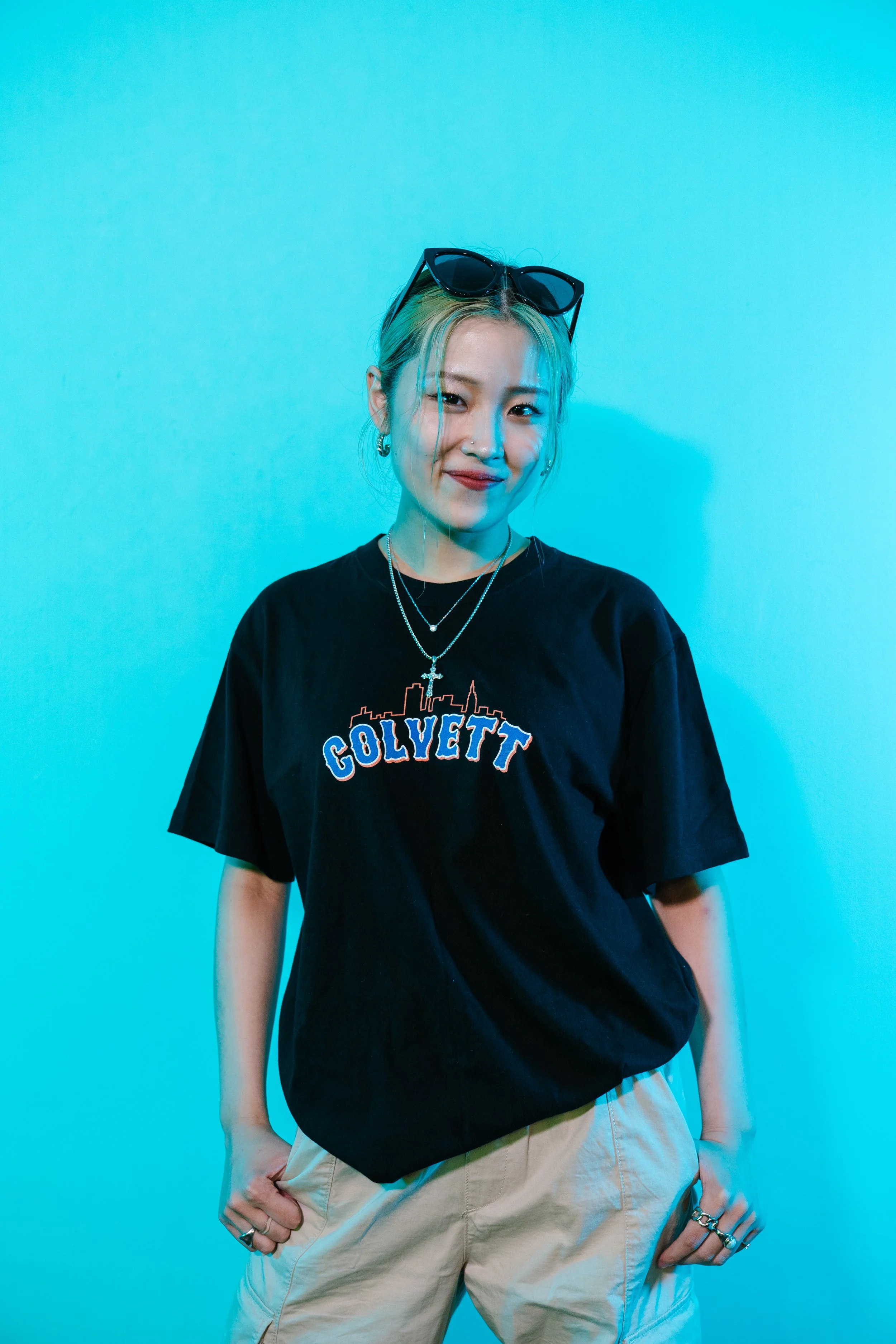 A young woman with blonde hair tied back, wearing black sunglasses on her head, a black t-shirt with colorful text and graphics, layered necklaces, earrings, and multiple rings, standing against a bright blue background with her hand in her pocket.