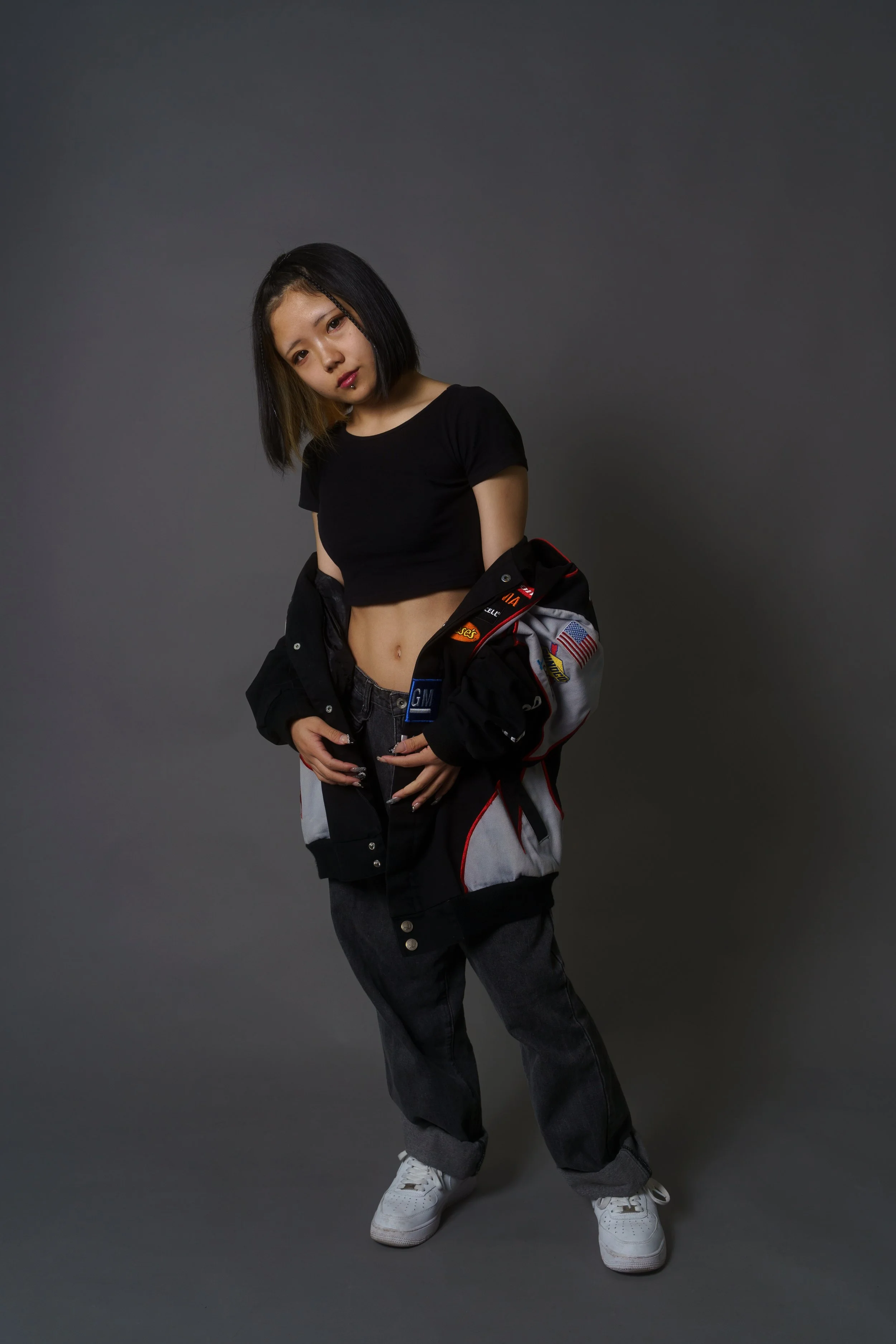Young woman with shoulder-length black hair, wearing a black crop top and baggy black pants with white sneakers, standing against a plain gray background.
