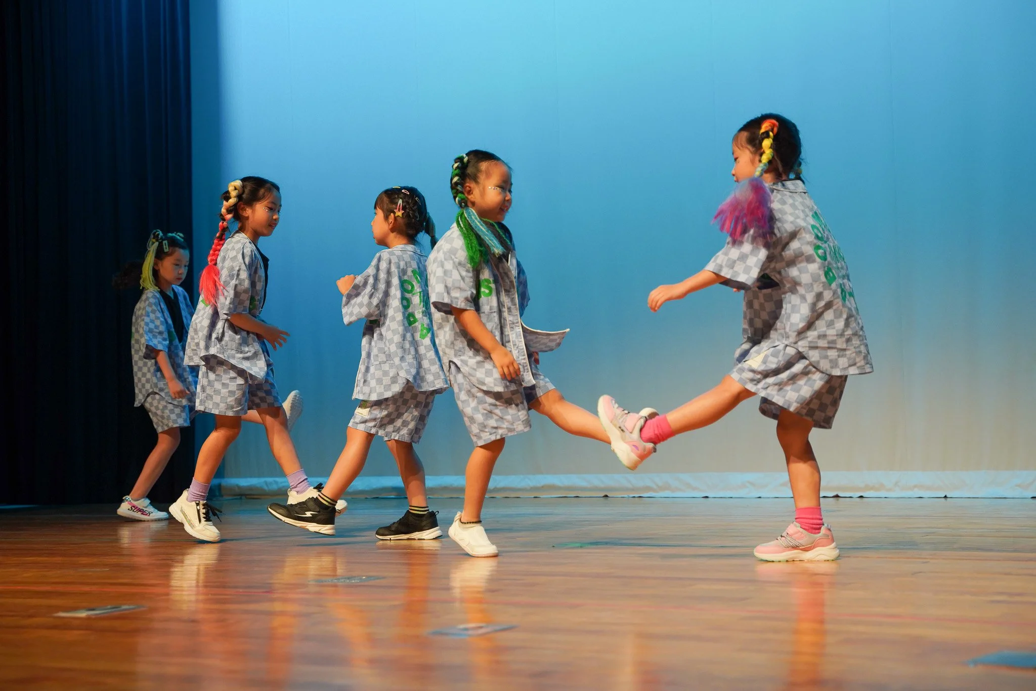 A group of young children dressed in gray checkered outfits participating in a dance or activity on stage, with two girls holding hands and leaping while others wait in line, colorful hair accessories, sneakers, and a blue backdrop.