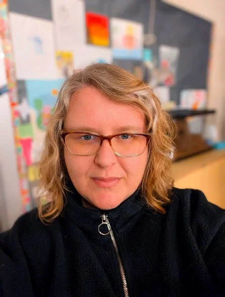 A close-up portrait of Danielle, the founder of Blue Horizon Studio. She is an 18-year veteran educator with blonde wavy hair and glasses, pictured in a warm, supportive studio environment with a colorful display of student work in the background.
