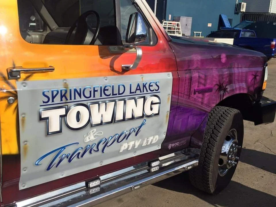 A pickup truck with a colorful paint job featuring pink, purple, and orange hues, advertising 'Springfield Lakes Towing & Transport PTY LTD' on the door. The truck has black tires and appears to be parked in an outdoor lot.