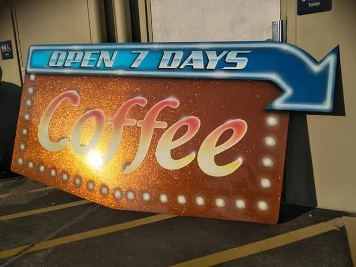 A brightly lit sign advertising a coffee shop that is open 7 days a week, with a large arrow pointing to the right.