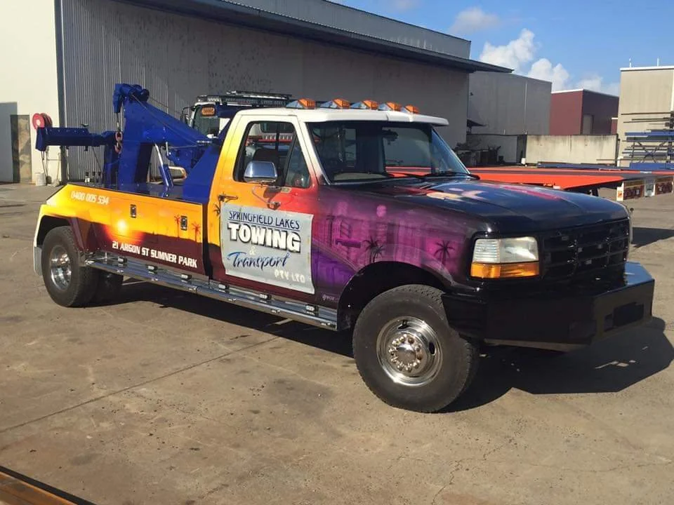 A tow truck with colorful sunset and palm tree artwork on the side, parked outdoors on a paved surface, with a warehouse in the background.
