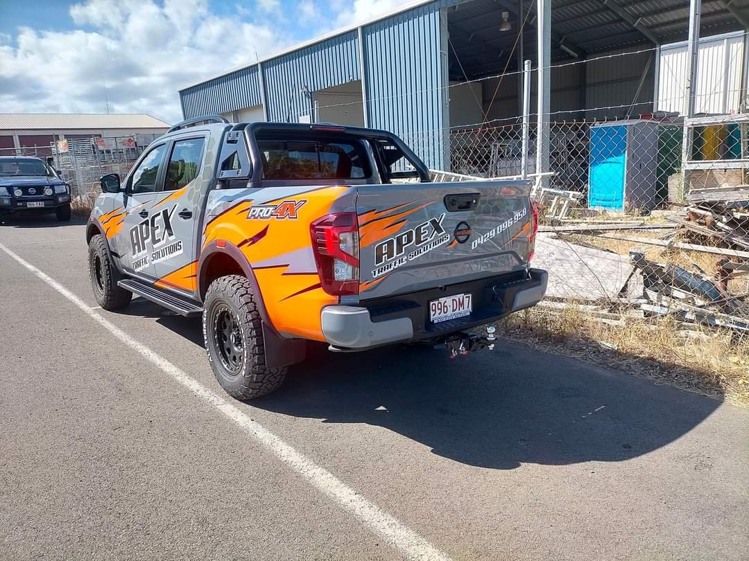 A pickup truck with baseball-sized tires and a colorful graphic wrap with the words 'APEX Traffiic Solutions' and 'Pro4X' parked next to a blue industrial building and a fenced area with various objects.