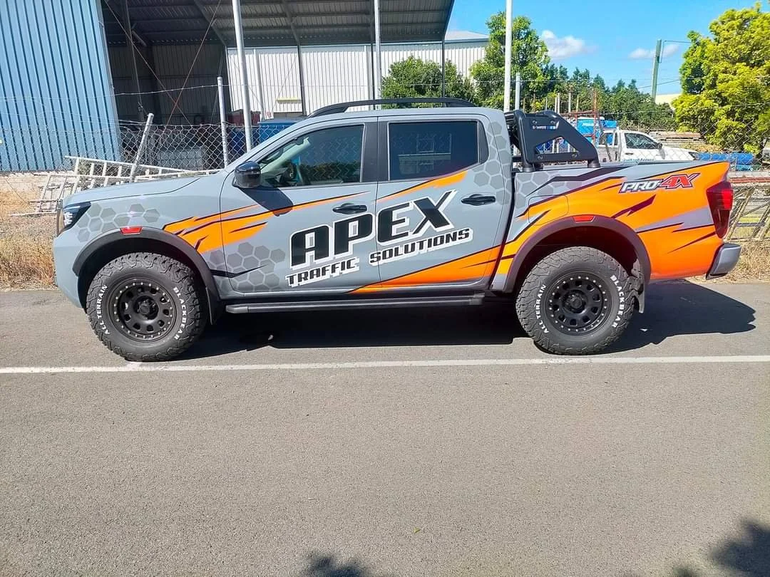A gray pickup truck with a graphic design that includes orange flames and honeycomb patterns. The truck has the text 'APEX Traffic Solutions' and 'Pro-X' on it, parked on a paved street with an industrial building, fence, and trees in the background.