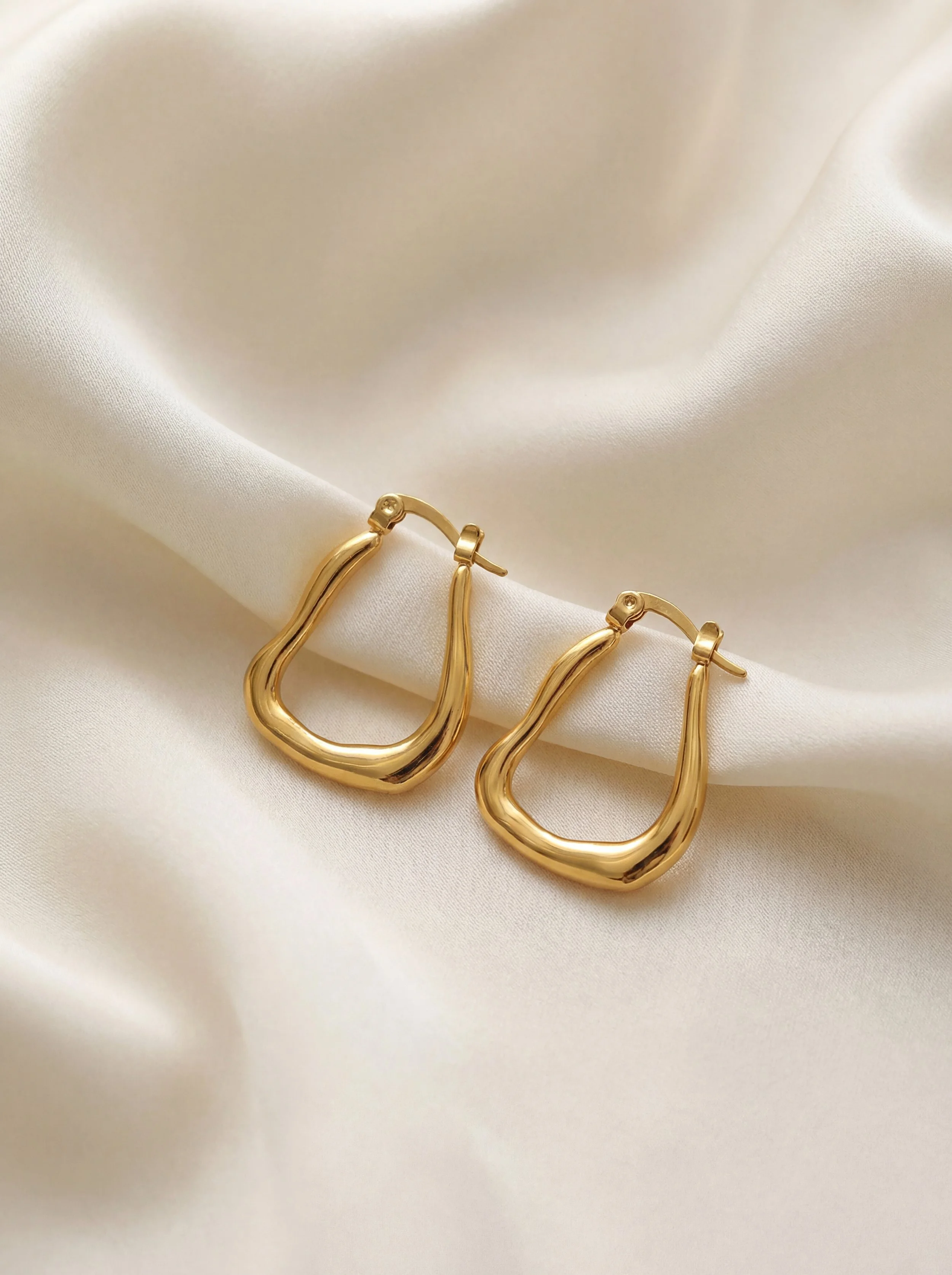 A pair of gold earrings with a wavy, open hoop design resting on ivory satin fabric.