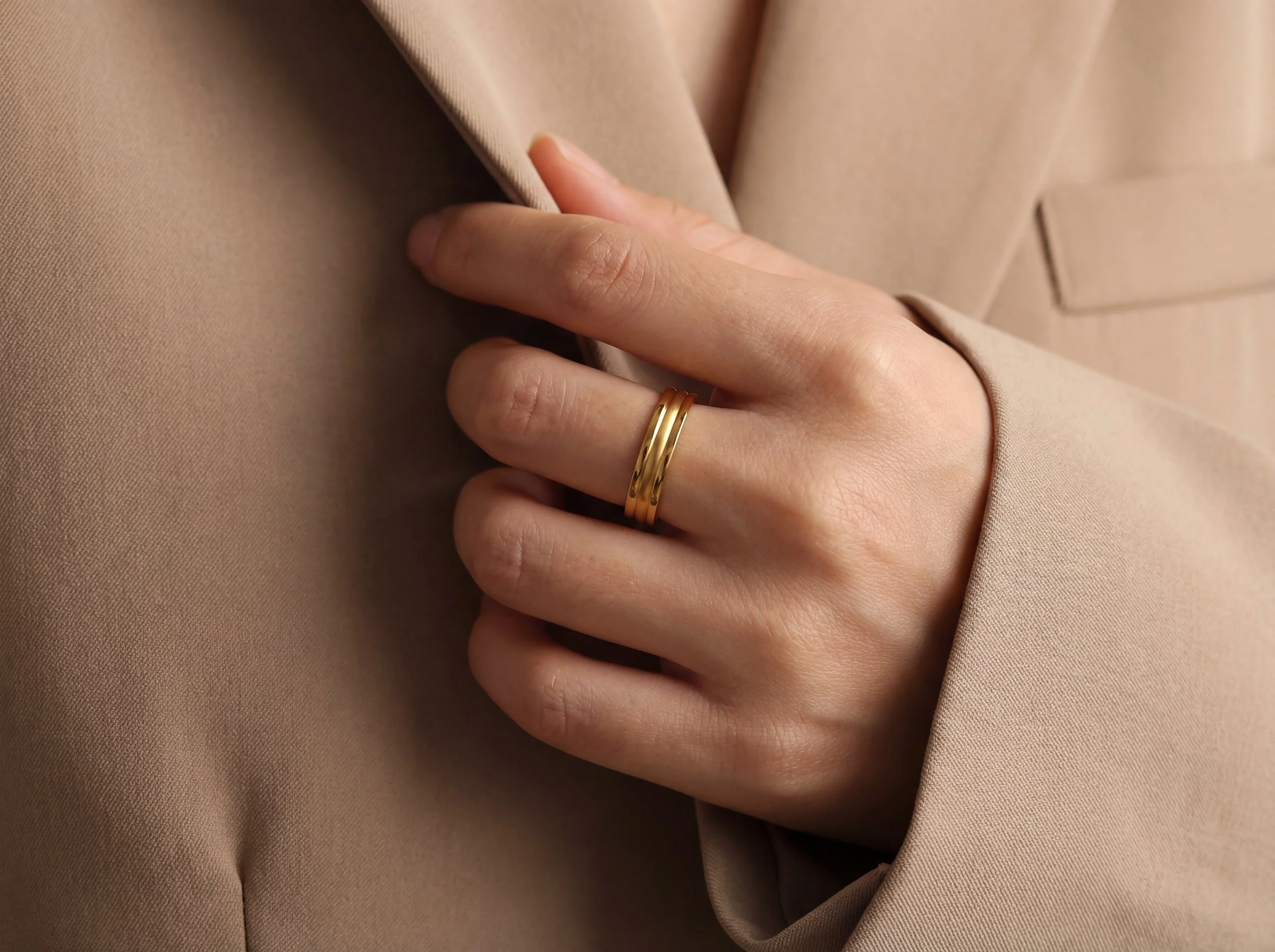 Close-up of a person's hand with a gold ring on the ring finger, touching a beige blazer.