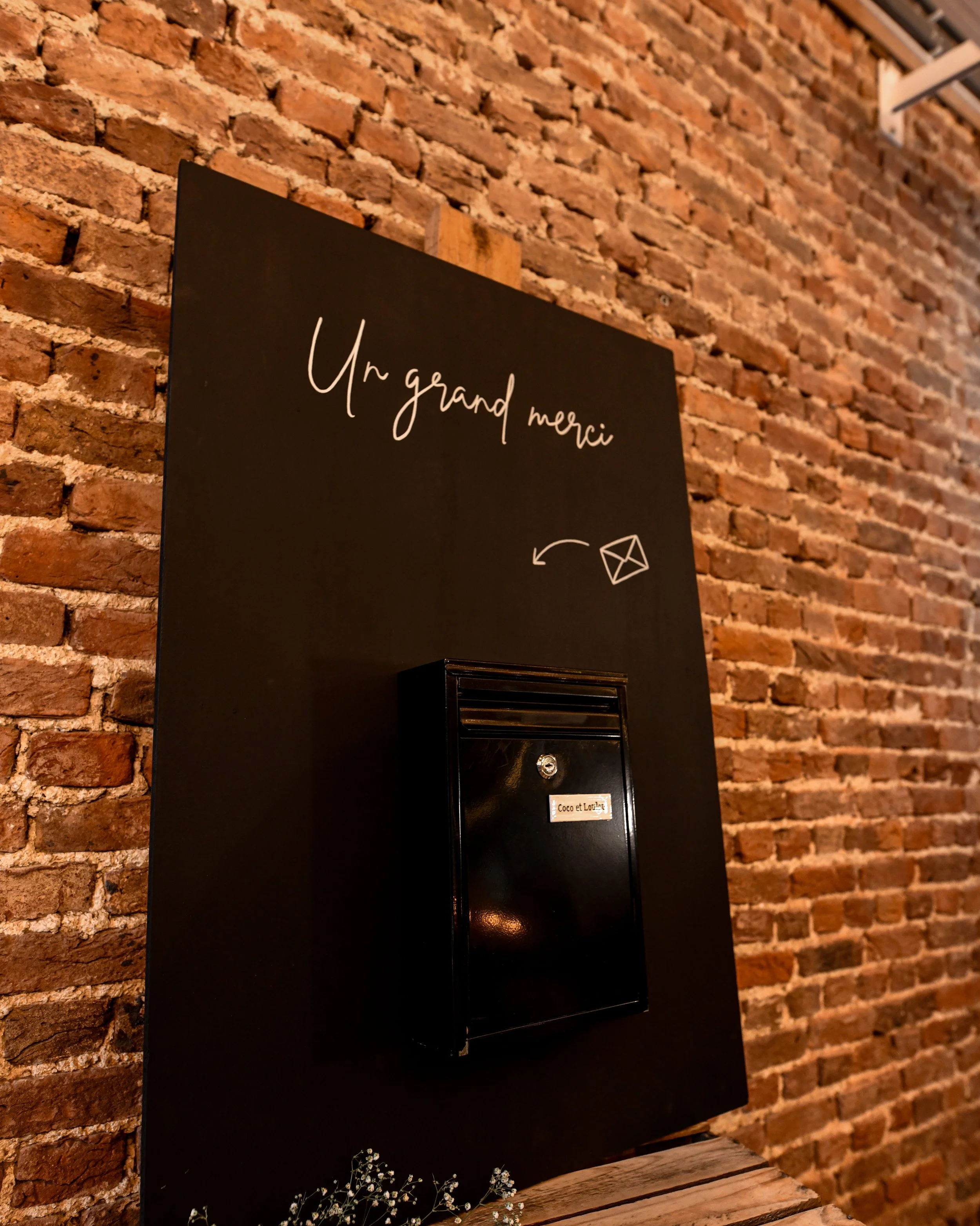 Une boîte aux lettres noire fixée à un mur en briques rouges avec une inscription en français "Un grand merci" et un symbole de courrier, décorée avec des petites fleurs blanches en bas.