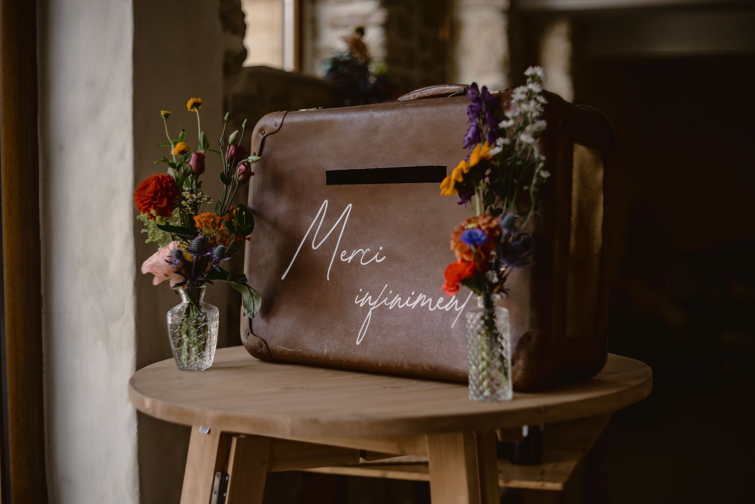 Un panneau en cuir avec le mot « Merci » écrit en cursive blanche, entouré de bouquets de fleurs dans des vases en verre, placé sur une table en bois