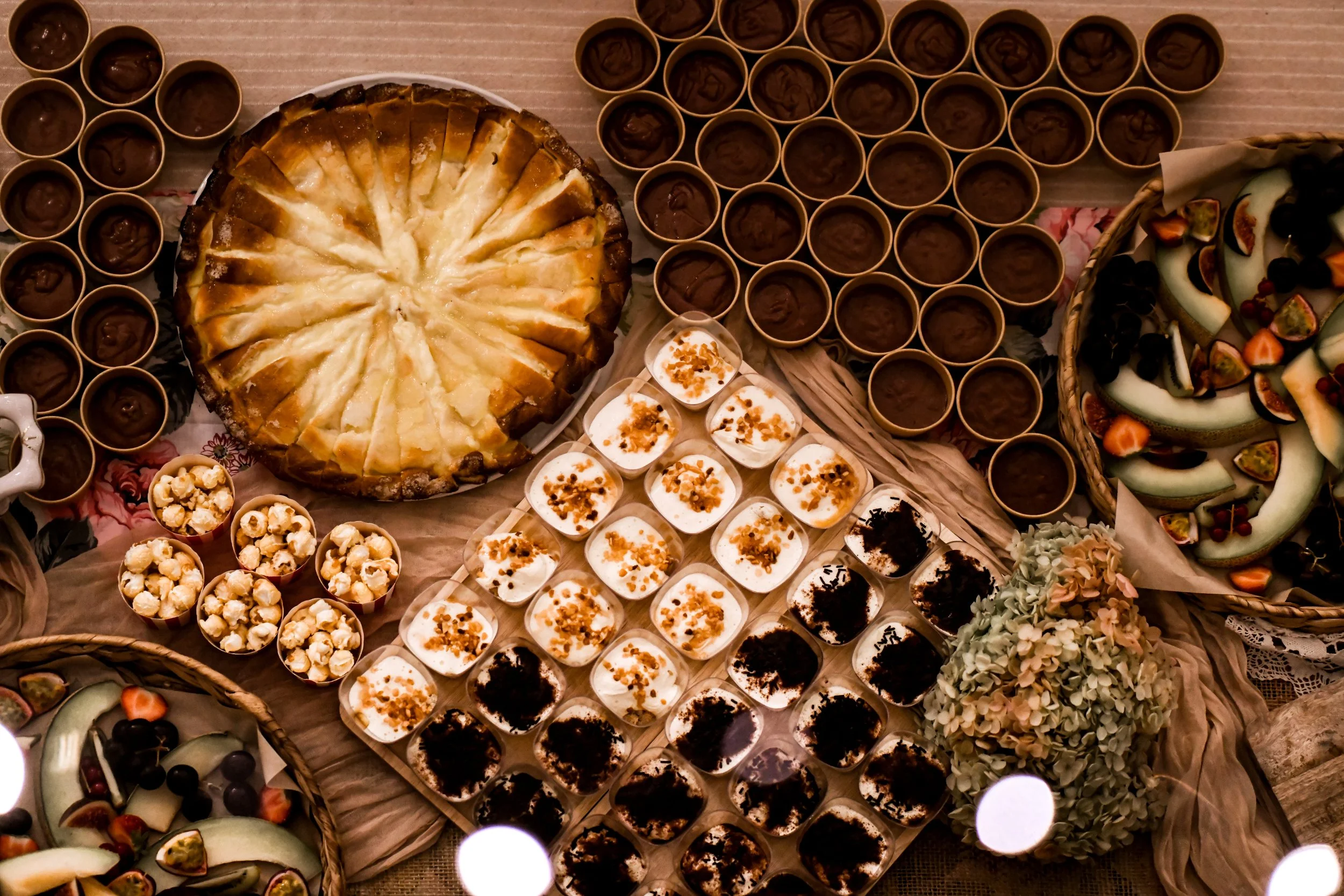 Table recouverte de desserts variés comme un cheesecake, des petits pot de crème, des cupcakes, un assortiment de fruits, et des chocolats en petits pots.