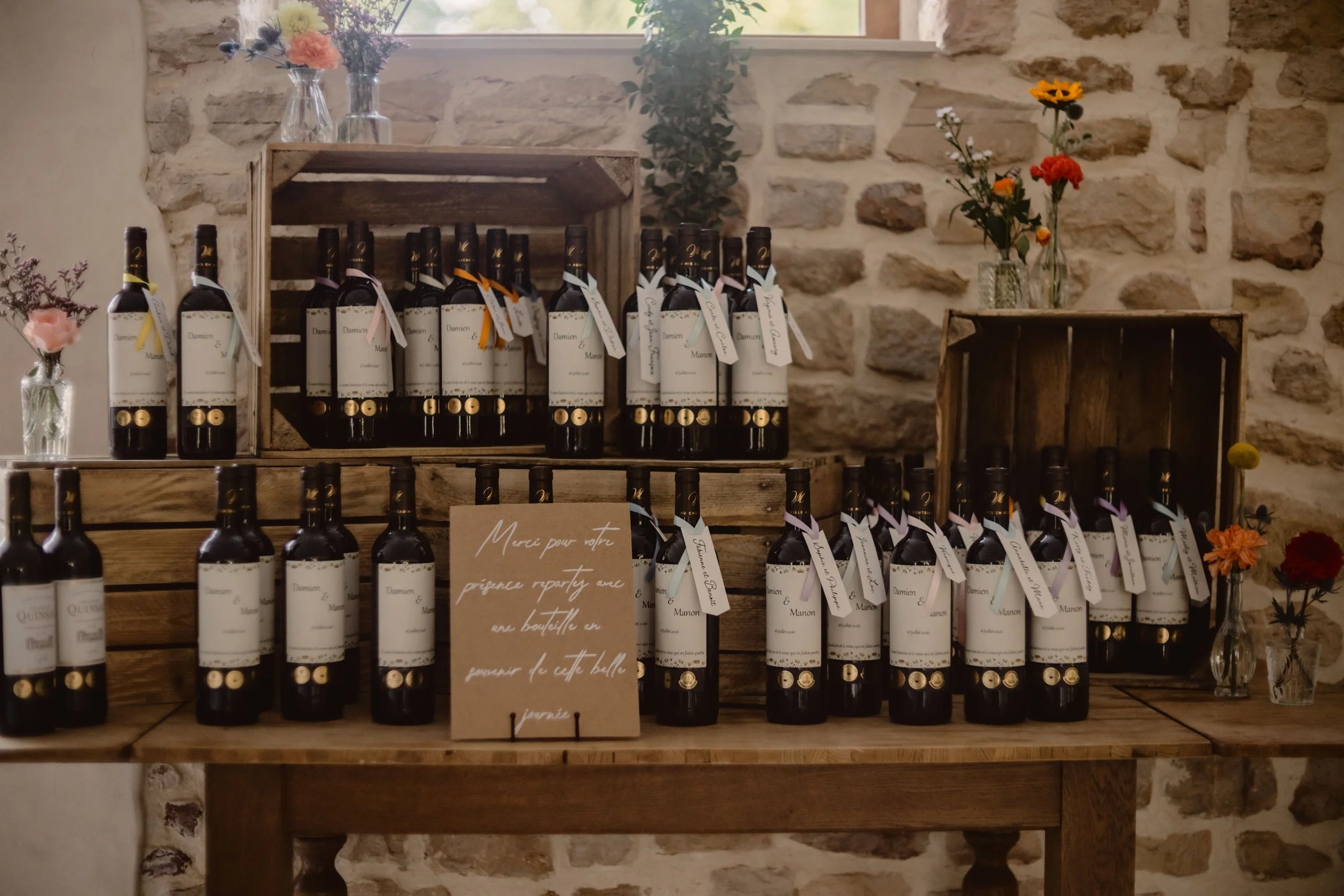 Bouteilles de vin alignées sur une table en bois, décorées de petits rubans de couleur, avec des fleurs dans des vases et un mur en pierre en arrière-plan.