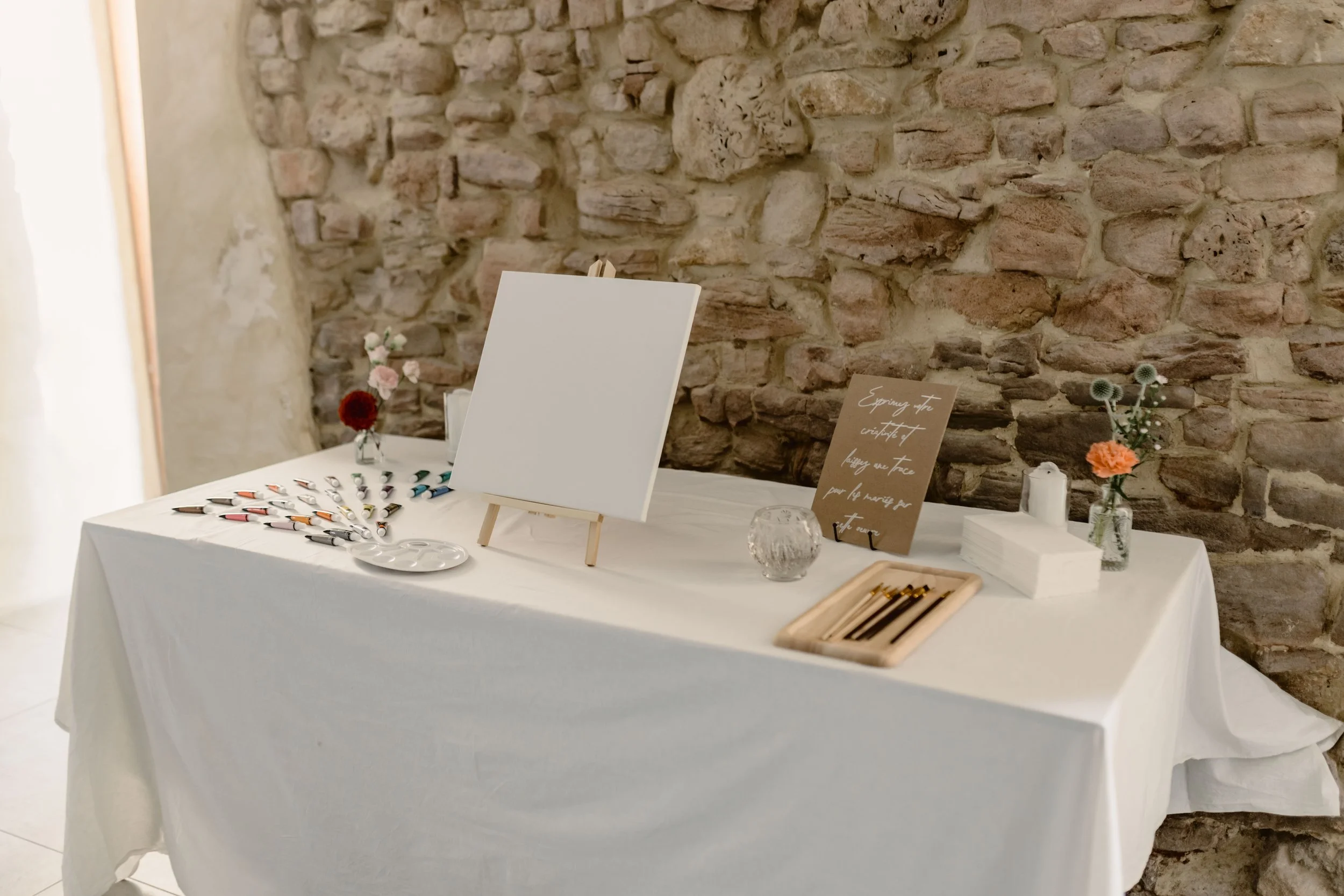 Table décorée avec des fleurs, stylos et pinceaux, contre un mur en pierre, avec une toile blanche et une carte avec un message.