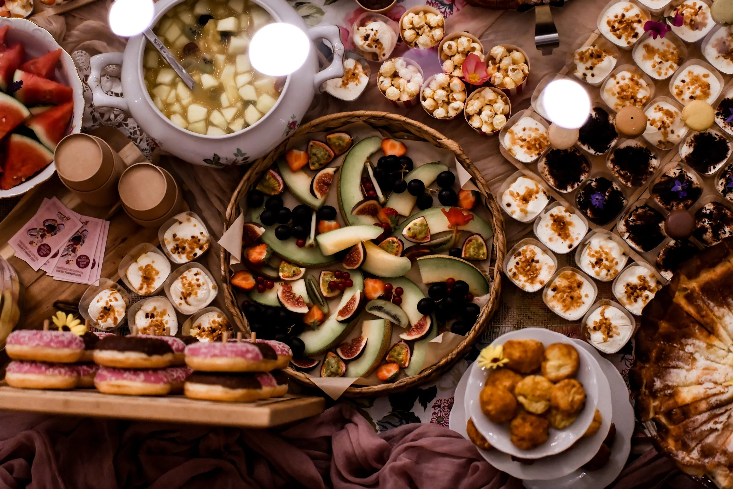 Une variété de desserts et de fruits présentés sur une table, incluant des cupcakes, des tranches de pastèque, un plateau de fruits frais comme melon, figues, raisins noirs, et d'autres petits gâteaux et friandises.