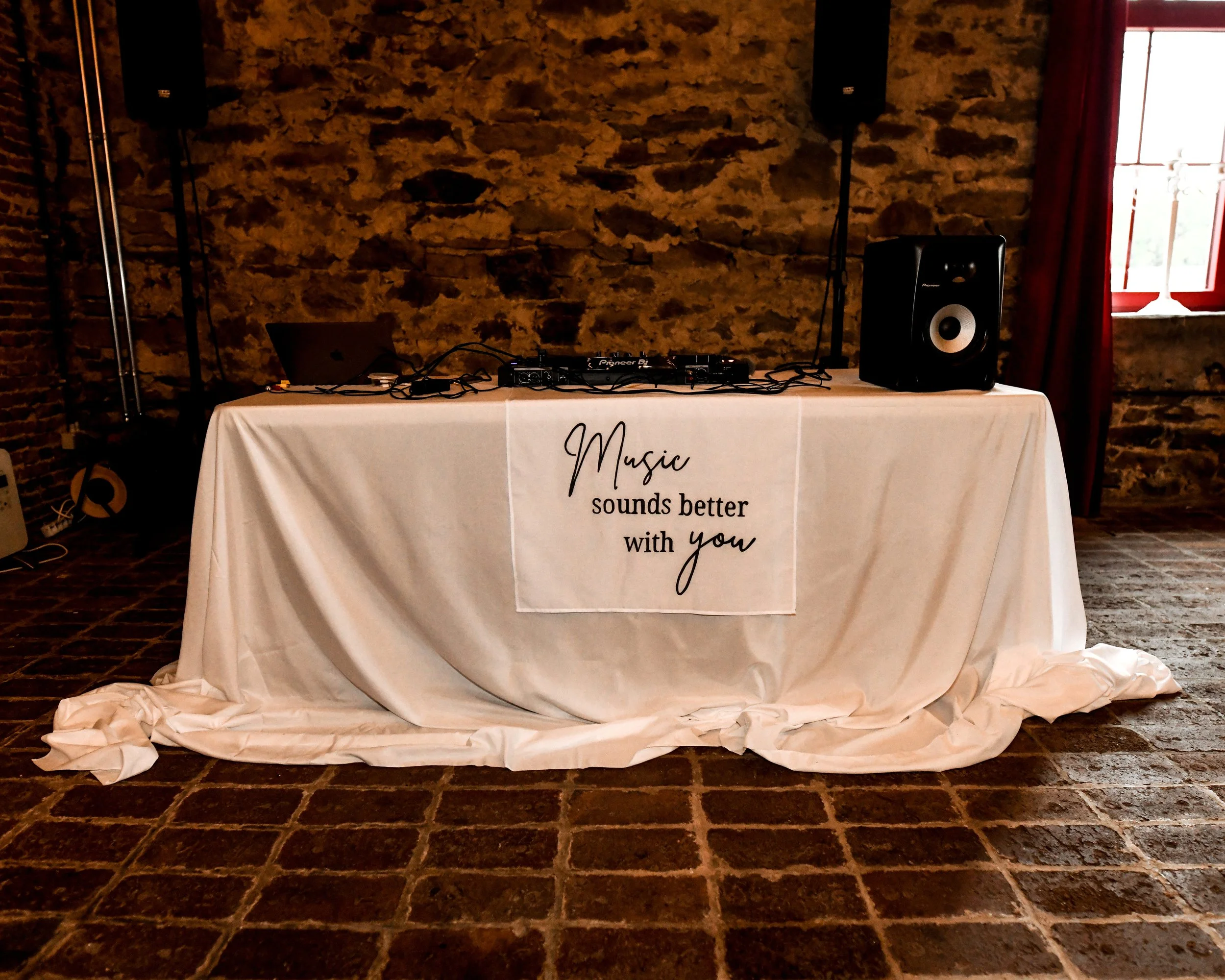 Tableau avec une bannière blanche portant l'inscription "Music sounds better with you", un ordinateur portable, un lecteur audio, haut-parleurs, et des câbles, dans une pièce avec un mur en pierre et une fenêtre avec rideau rouge.