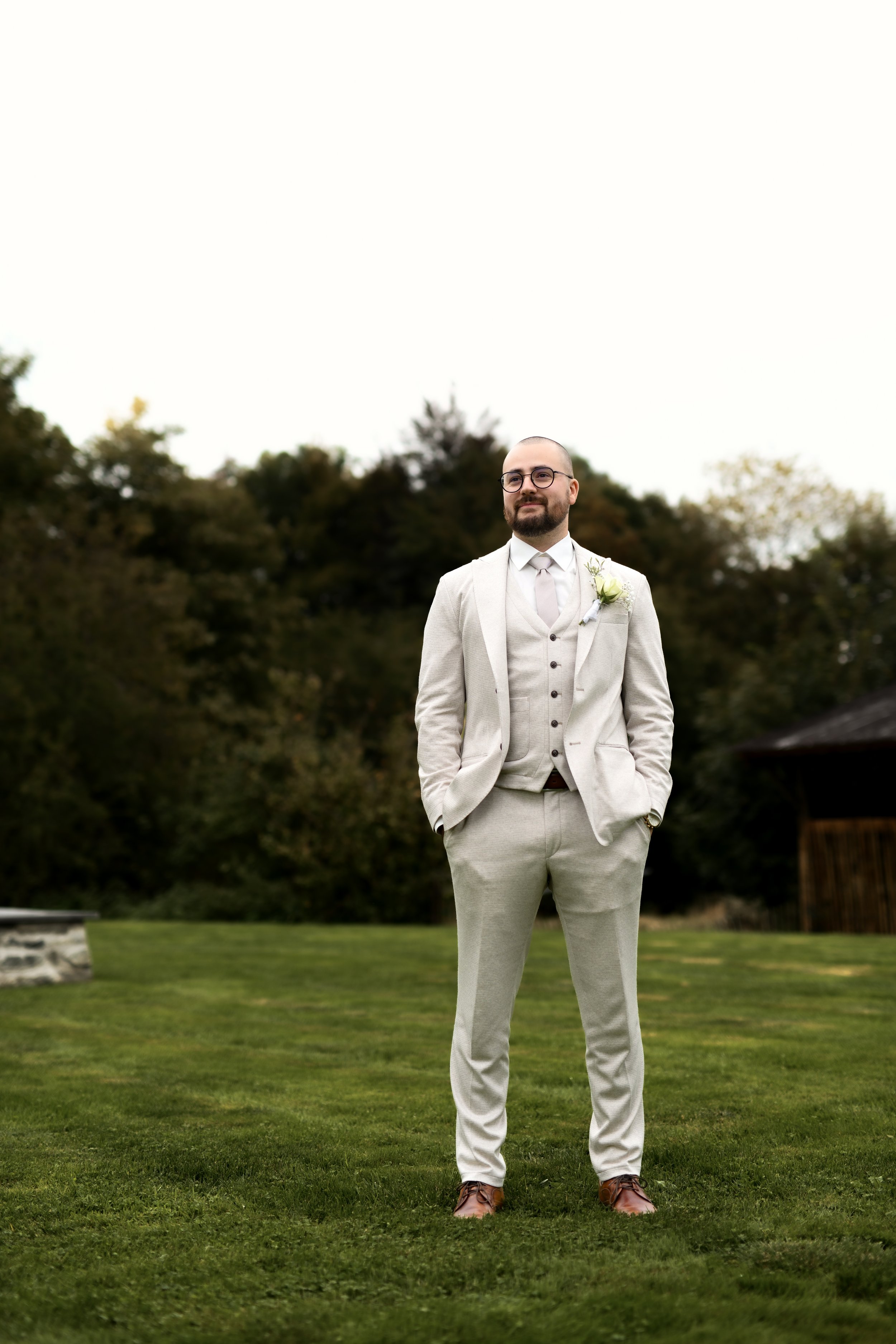 Homme en costume beige, portant une boutonnière, debout sur une pelouse verte avec des arbres en arrière-plan, ciel clair au-dessus.