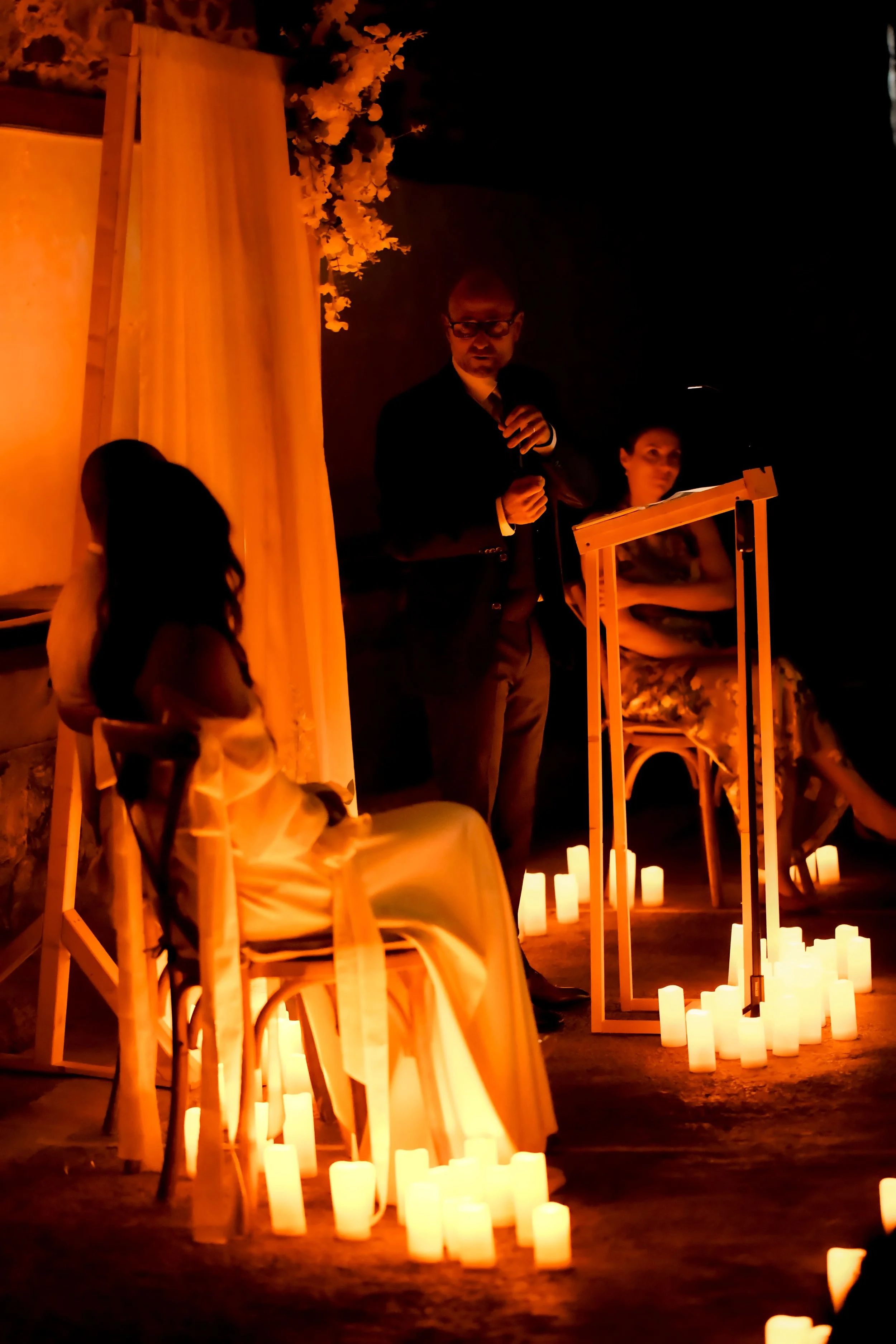 Une cérémonie ou un discours se déroule la nuit, avec des bougies ou des lumières tamisées, des chaises en bois, une personne debout parlant dans un micro, un cadre en bois, une femme assise à droite, et un décor simple avec des fleurs et des draperi
